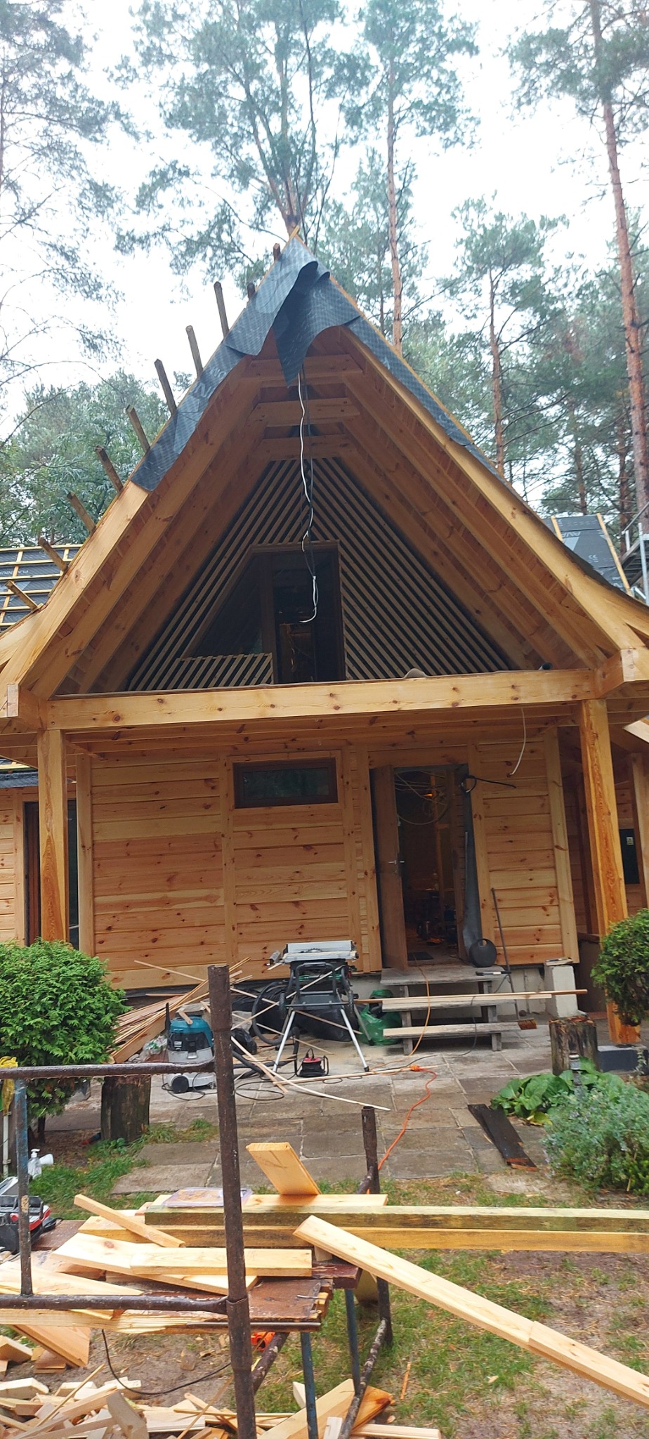 Budowa domu z drewnianych bali w lesie, widoczny etap montażu dachu z membraną, narzędzia budowlane i resztki materiałów na placu budowy.