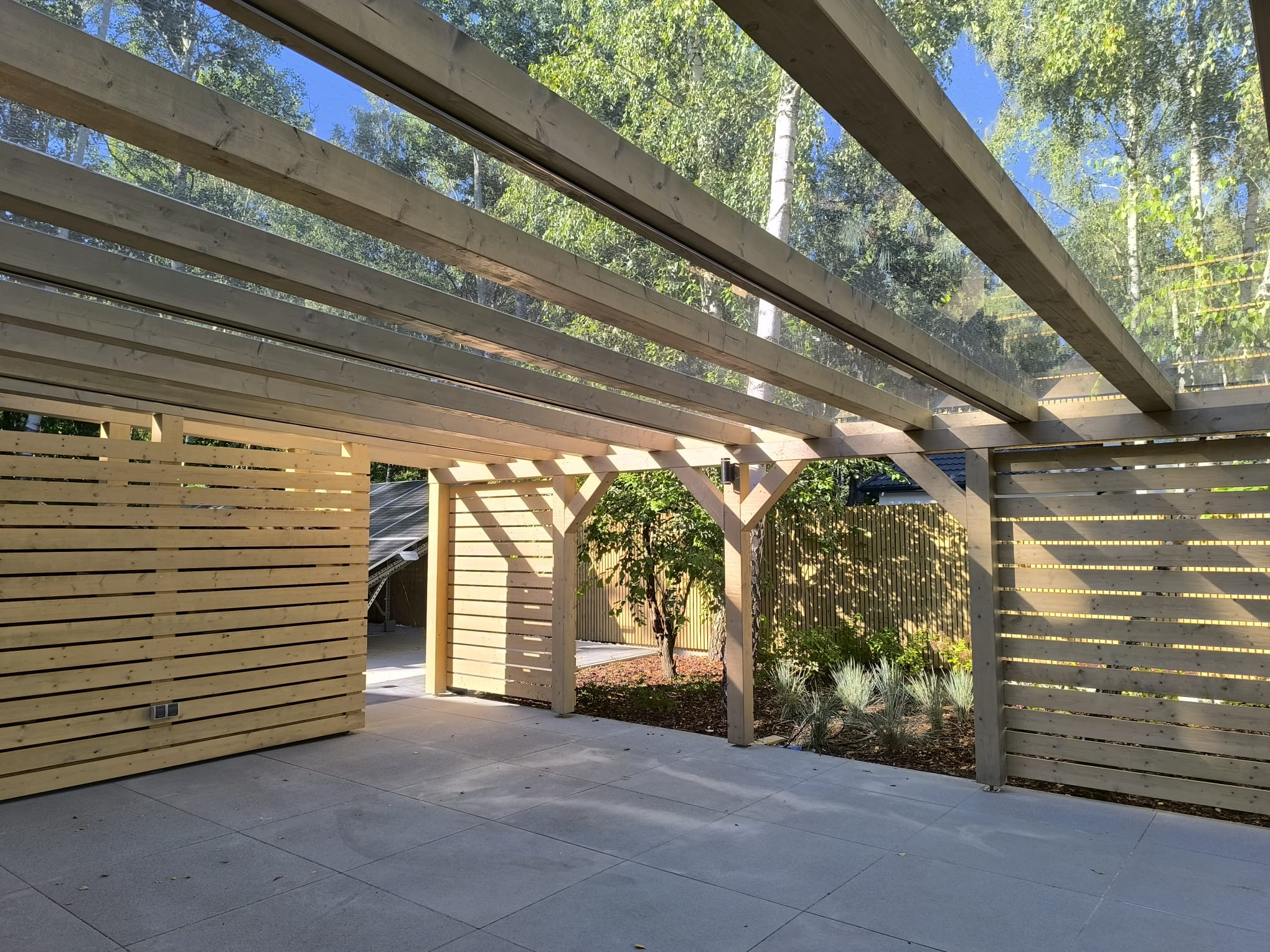 Drewniana konstrukcja z zadaszeniem, tworząca zadaszone patio z elementami ażurowej zabudowy, widoczne drzewa w tle.