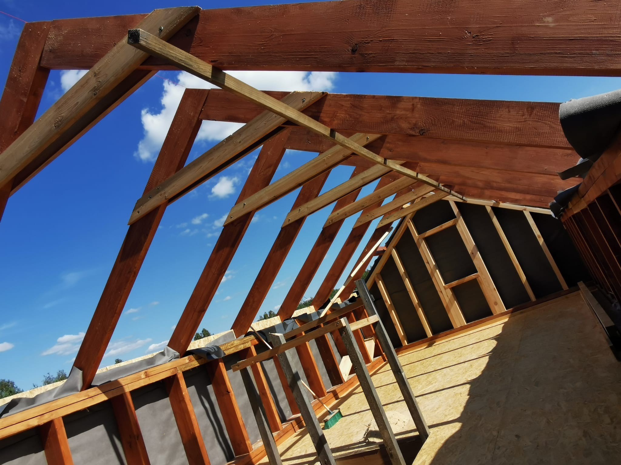 Wnętrze w trakcie budowy, widoczne drewniane elementy konstrukcyjne dachu i ścian, zabezpieczone czarną membraną, na tle błękitnego nieba z chmurami.