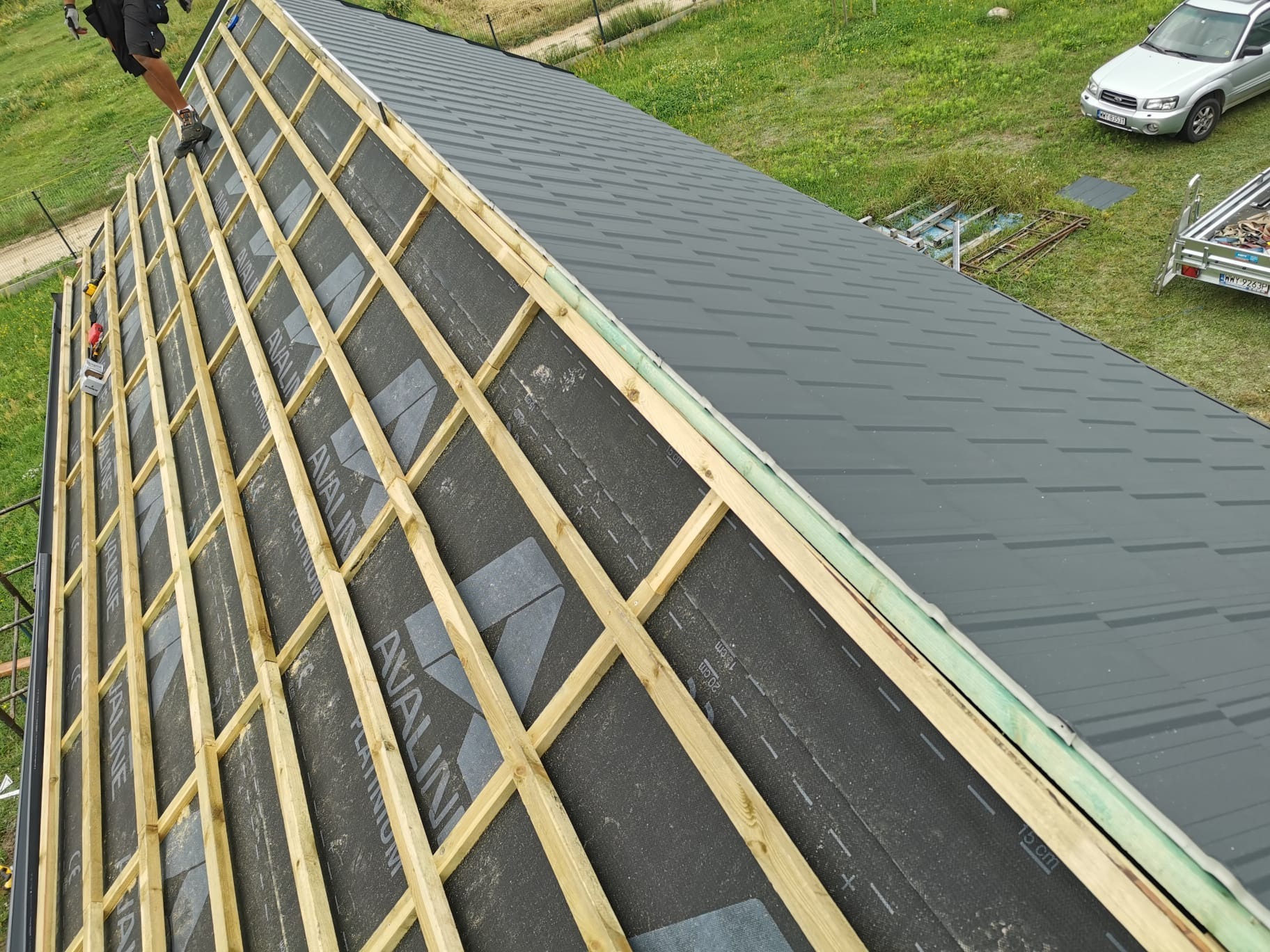 Montaż pokrycia dachu z blachy na drewnianej konstrukcji, widoczna membrana dachowa z nadrukiem, w tle zielona trawa i samochód osobowy z przyczepką.