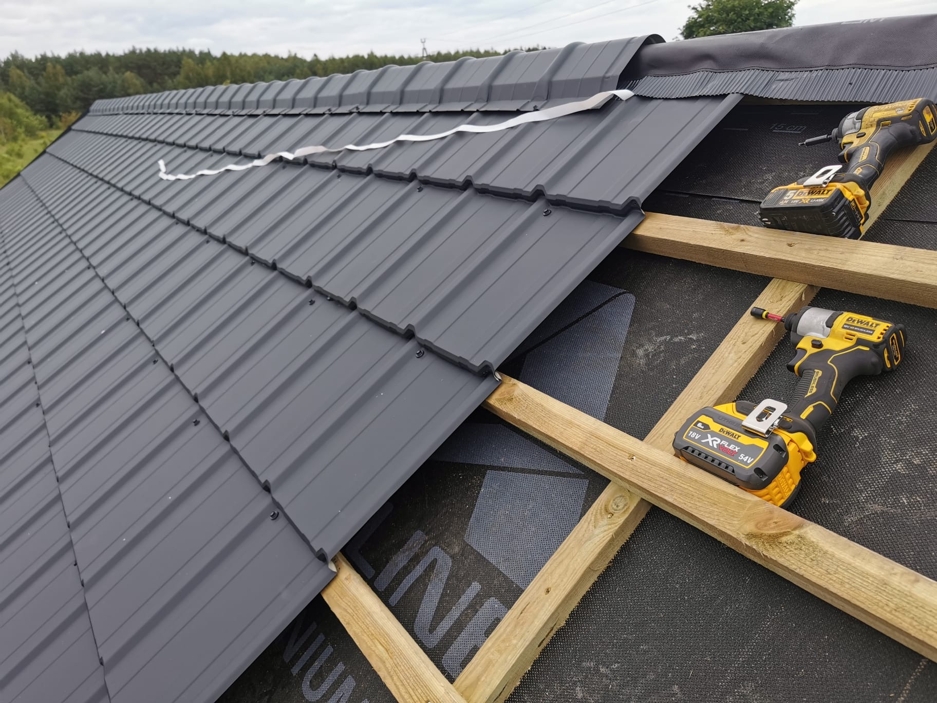 Montaż szarej blachodachówki na drewnianej konstrukcji dachu, widoczne dwa akumulatorowe wkrętaki DeWalt i fragment taśmy uszczelniającej.