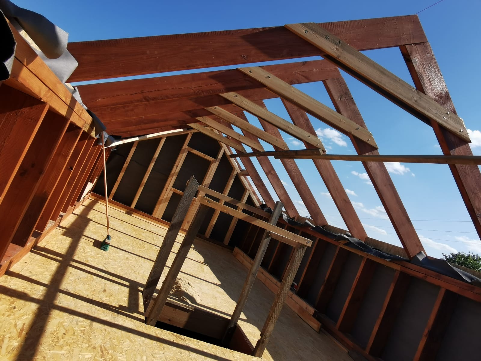 Wnętrze w trakcie budowy domu szkieletowego: widok na konstrukcję dachu z belek, płyty OSB na podłodze i czarną membranę na ścianach szczytowych, z fragmentem nieba widocznym przez otwory...