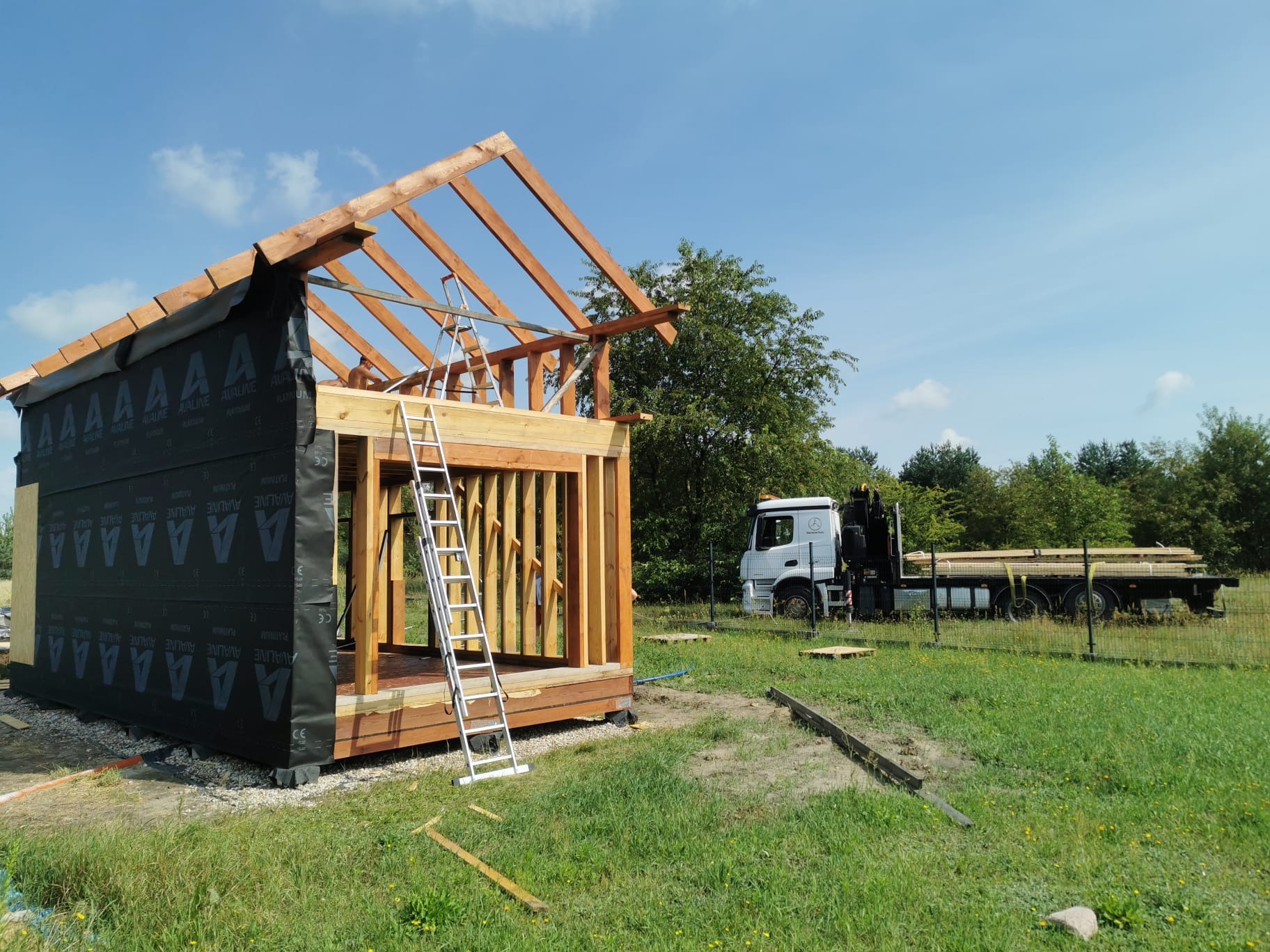 Wznoszenie konstrukcji drewnianej budynku w technologii szkieletowej z widoczną membraną izolacyjną, drabina oparta o konstrukcję dachu, w tle ciężarówka z transportem drewna na działce z zieloną...