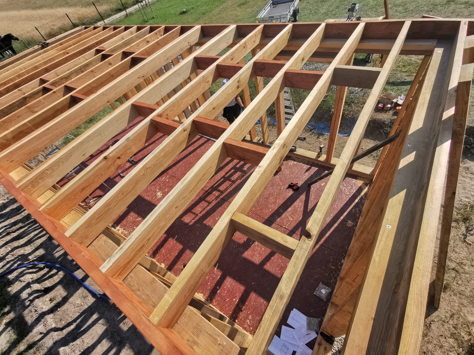 Konstrukcja szkieletowa domu w trakcie budowy w Białebłoto-Kobyla, widok z góry na drewniane belki i czerwone płyty podłogowe, częściowo zacienione.