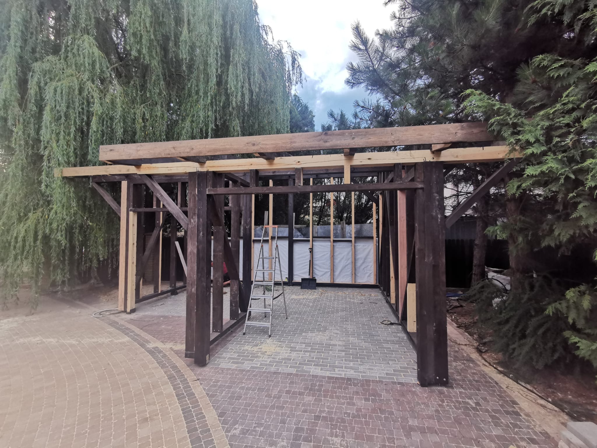 Konstrukcja drewniana z widoczną izolacją, drabina aluminiowa oparta o ścianę, kostka brukowa na podłożu, otoczenie drzew i krzewów.