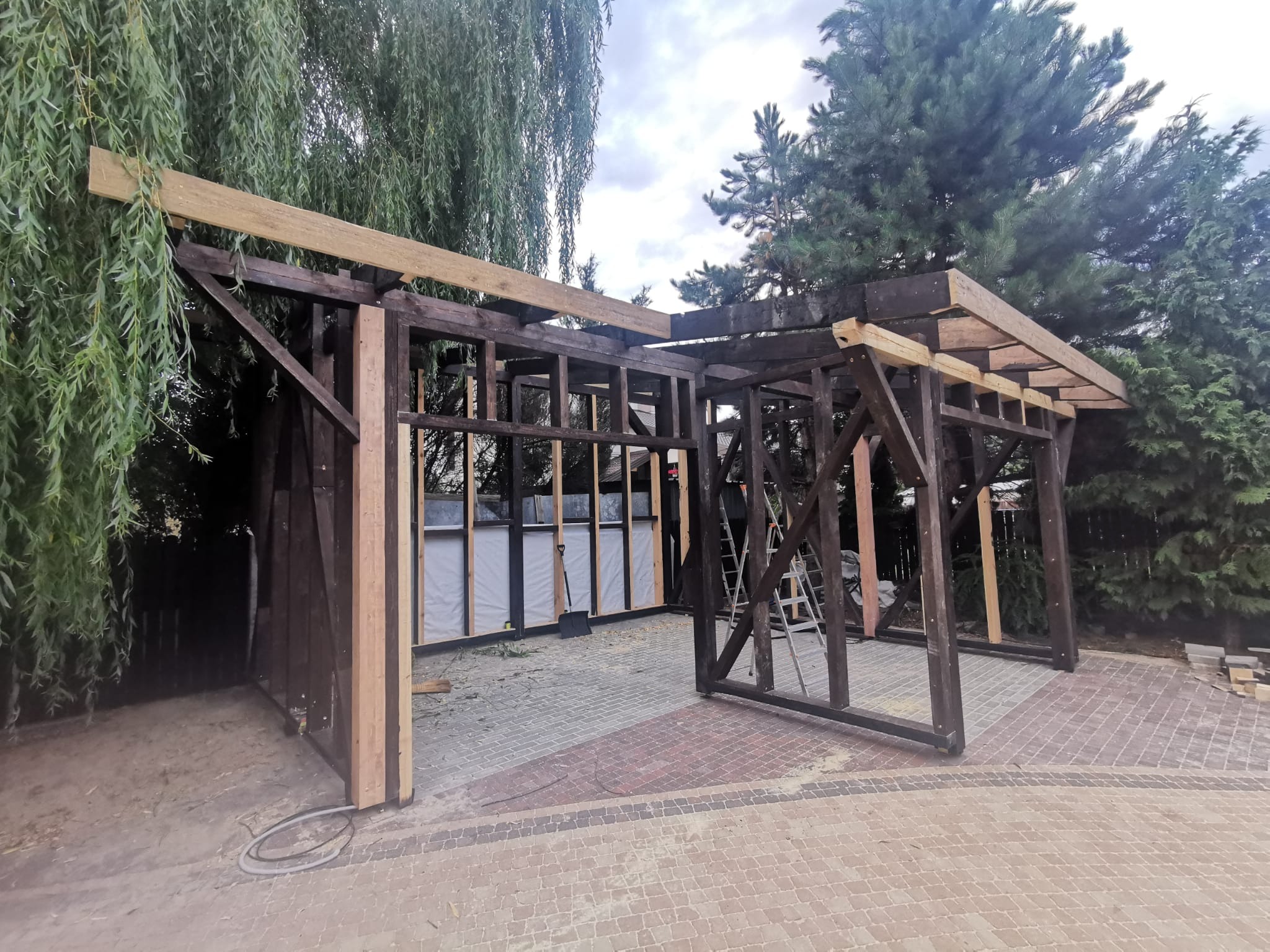Konstrukcja drewniana w trakcie budowy, widoczne elementy szkieletu, częściowo obite płytami, na zewnątrz kostka brukowa i zieleń