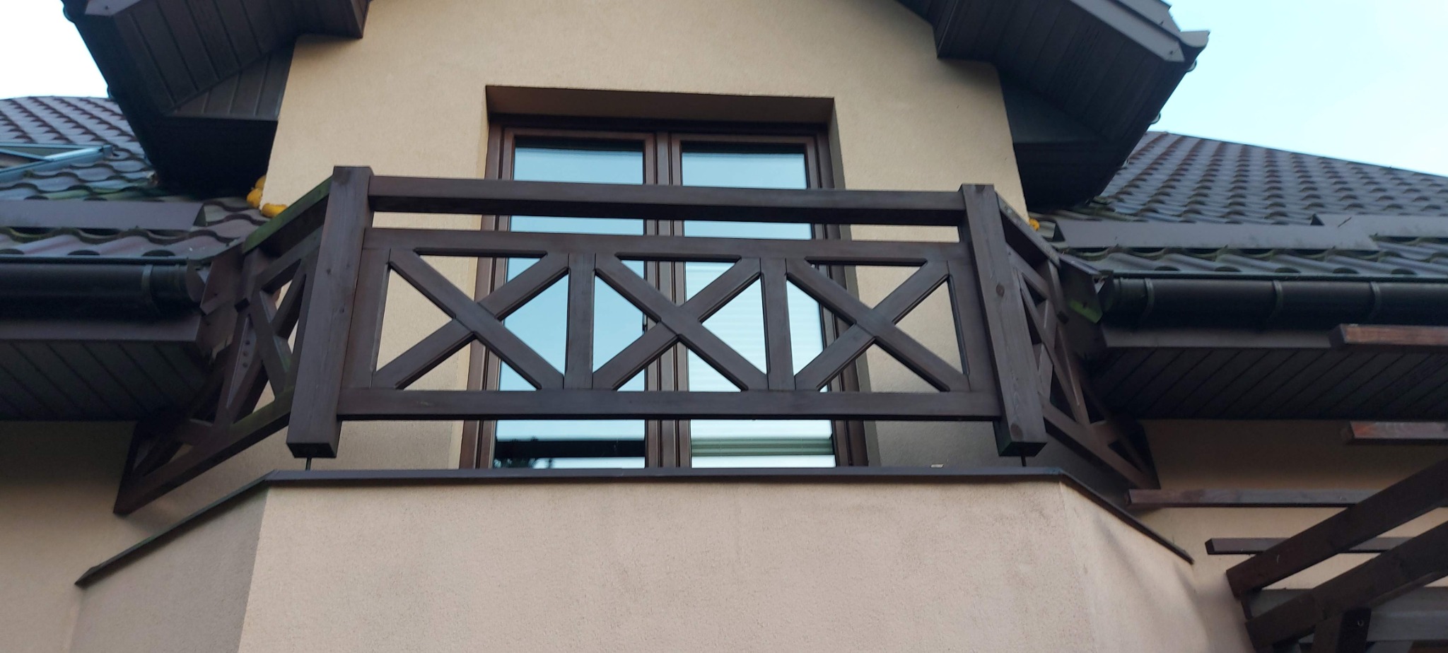 Fragment elewacji domu z beżowym tynkiem, balkon z ciemnobrązdką drewnianą balustradą o wzorze w krzyże, okno z odbijającym się niebem i fragment dachu z brązową dachówką.