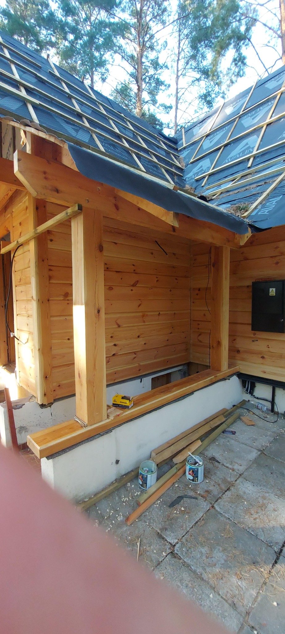 Konstrukcja z bali w trakcie budowy, widoczne drewniane ściany, elementy dachu z membraną, narzędzia i puszki z farbą na posadzce, otoczenie leśne.