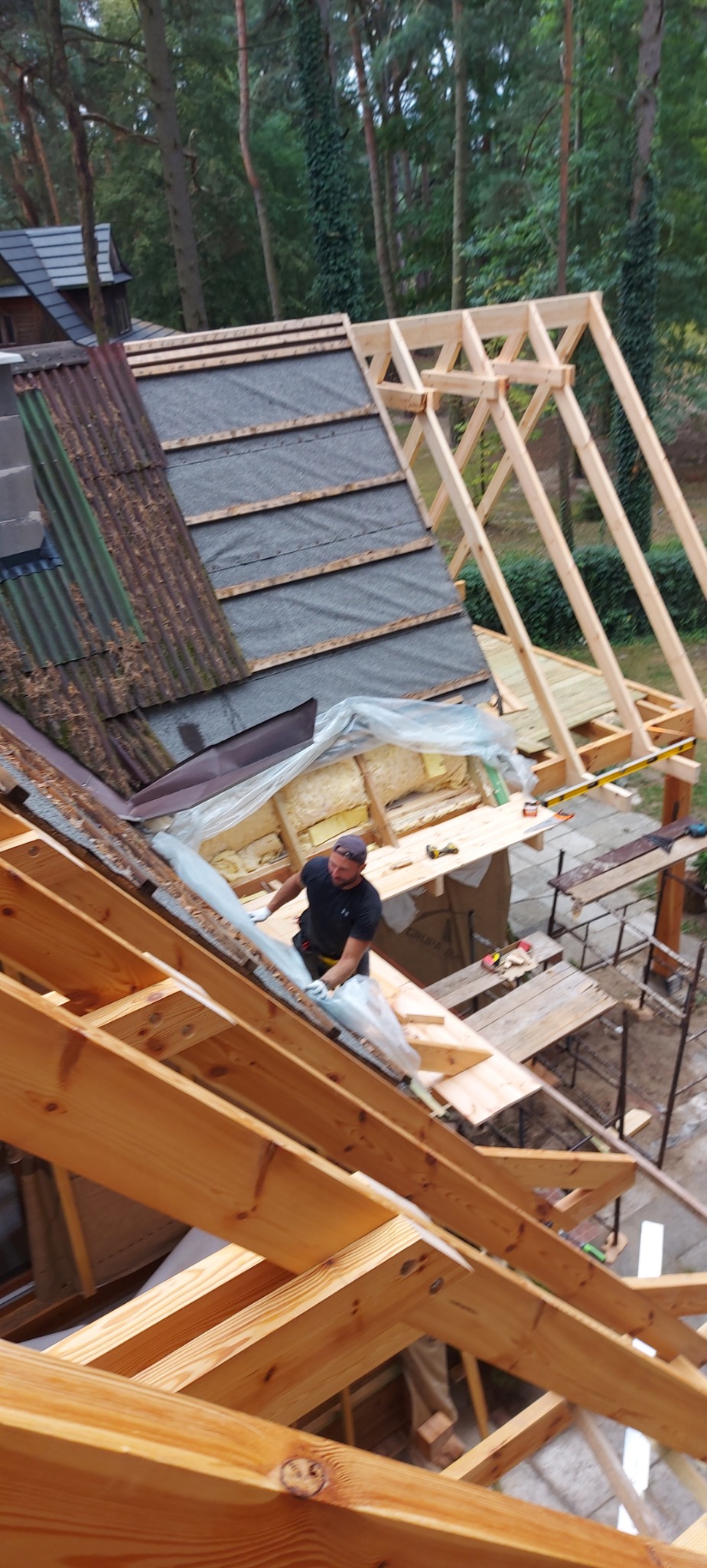 Mężczyzna w czarnej koszulce i czapce pracuje nad konstrukcją dachu drewnianego domu w lesie, widoczne elementy izolacji i różne pokrycia dachowe.