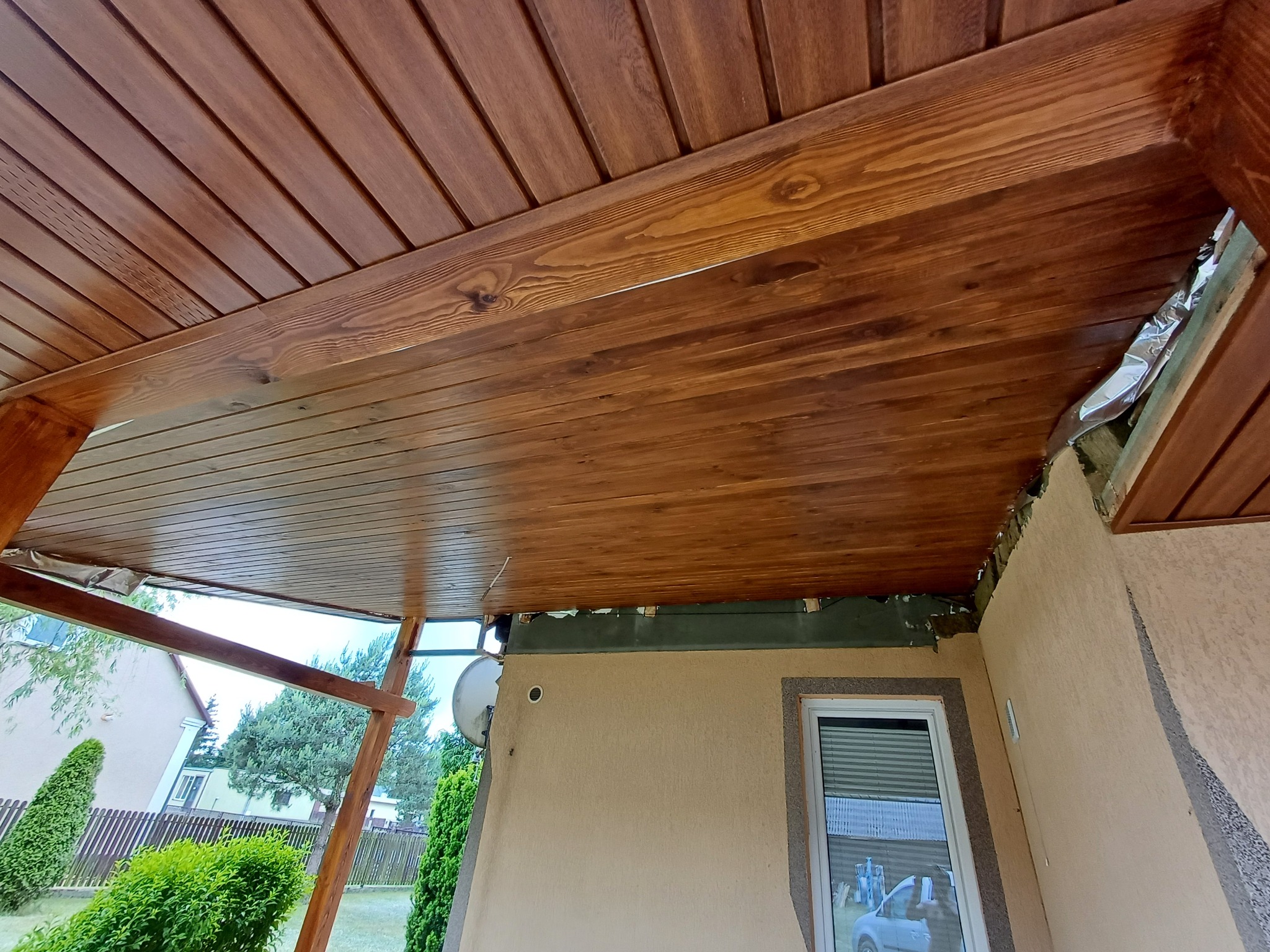 Drewniana podbitka dachowa z widoczną strukturą drewna, zamontowana na zewnątrz budynku, z widocznym fragmentem elewacji i ogrodu w tle.