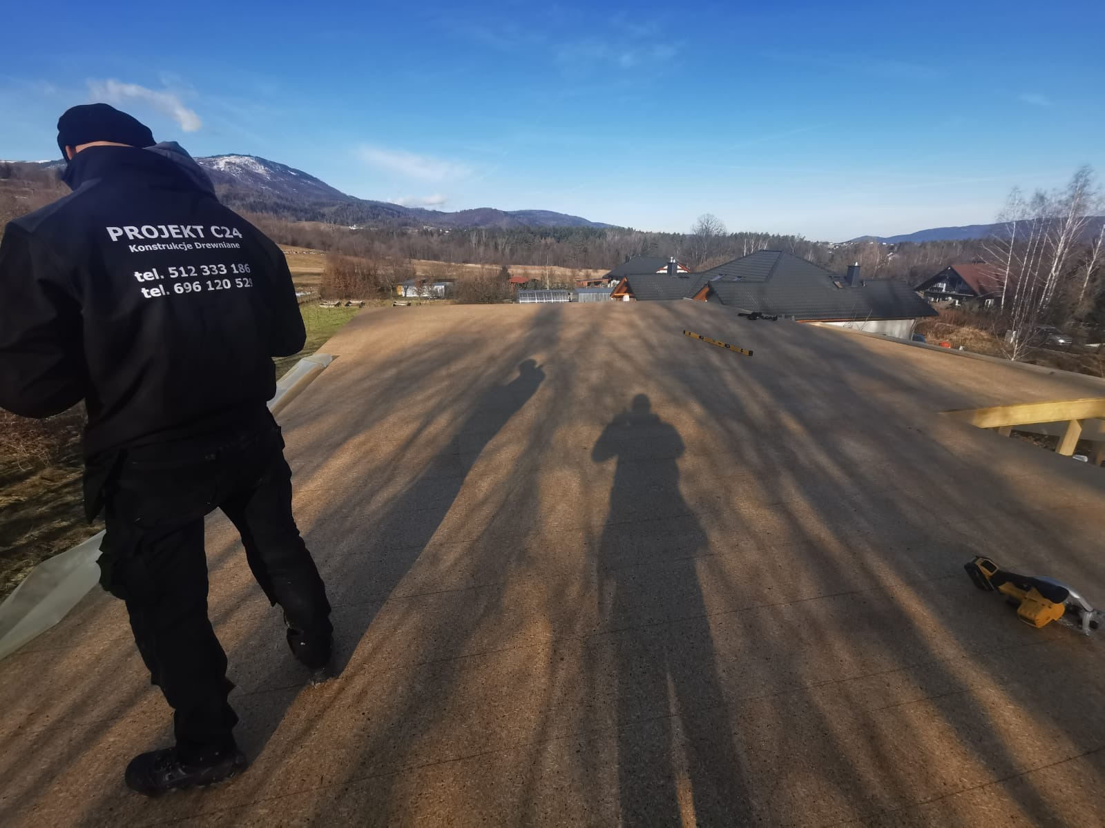 Ekipa budowlana na dachu z desek OSB, w tle góry i błękitne niebo, widoczna poziomica i narzędzie do cięcia.