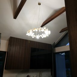 Fotovolt - Nowoczesny żyrandol w kształcie kwiatowego wieńca, oświetlający kuchnię z ciemnymi, drewnianymi szafkami i marmurowym blatem, widoczne drewniane belki stropowe.