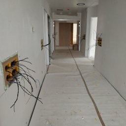Fotovolt - Korytarz w trakcie prac wykończeniowych, widoczne wystające przewody elektryczne z żółtymi puszkami instalacyjnymi w ścianach, zabezpieczona podłoga, surowy stan ścian.