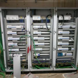 Rozdzielnia elektryczna składająca się z trzech szaf, widoczne liczne zabezpieczenia, przewody i szyny, w trakcie montażu, z elementami na podłodze.