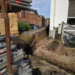 Fotovolt - Wykop w ziemi wzdłuż ściany budynku z widocznymi rurami i przewodami, elementy rusztowania w pierwszym planie.