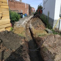 Fotovolt - Wykopany wąski rów w ziemi, prawdopodobnie pod kabel elektryczny, z widoczną stertą ziemi, stosem płyt izolacyjnych i fragmentem budynku w tle.