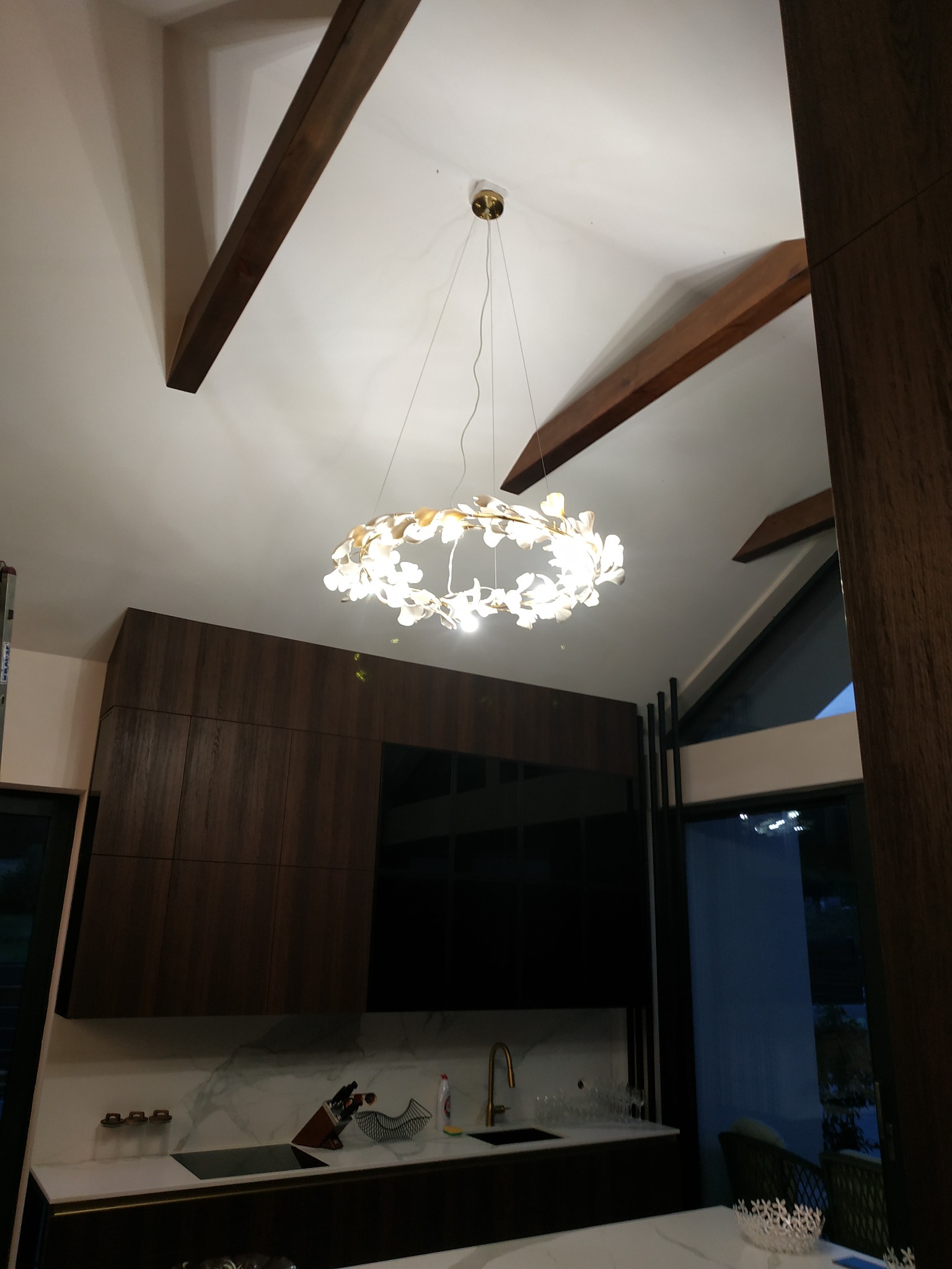 Nowoczesny żyrandol w kształcie kwiatowego wieńca, oświetlający kuchnię z ciemnymi, drewnianymi szafkami i marmurowym blatem, widoczne drewniane belki stropowe.