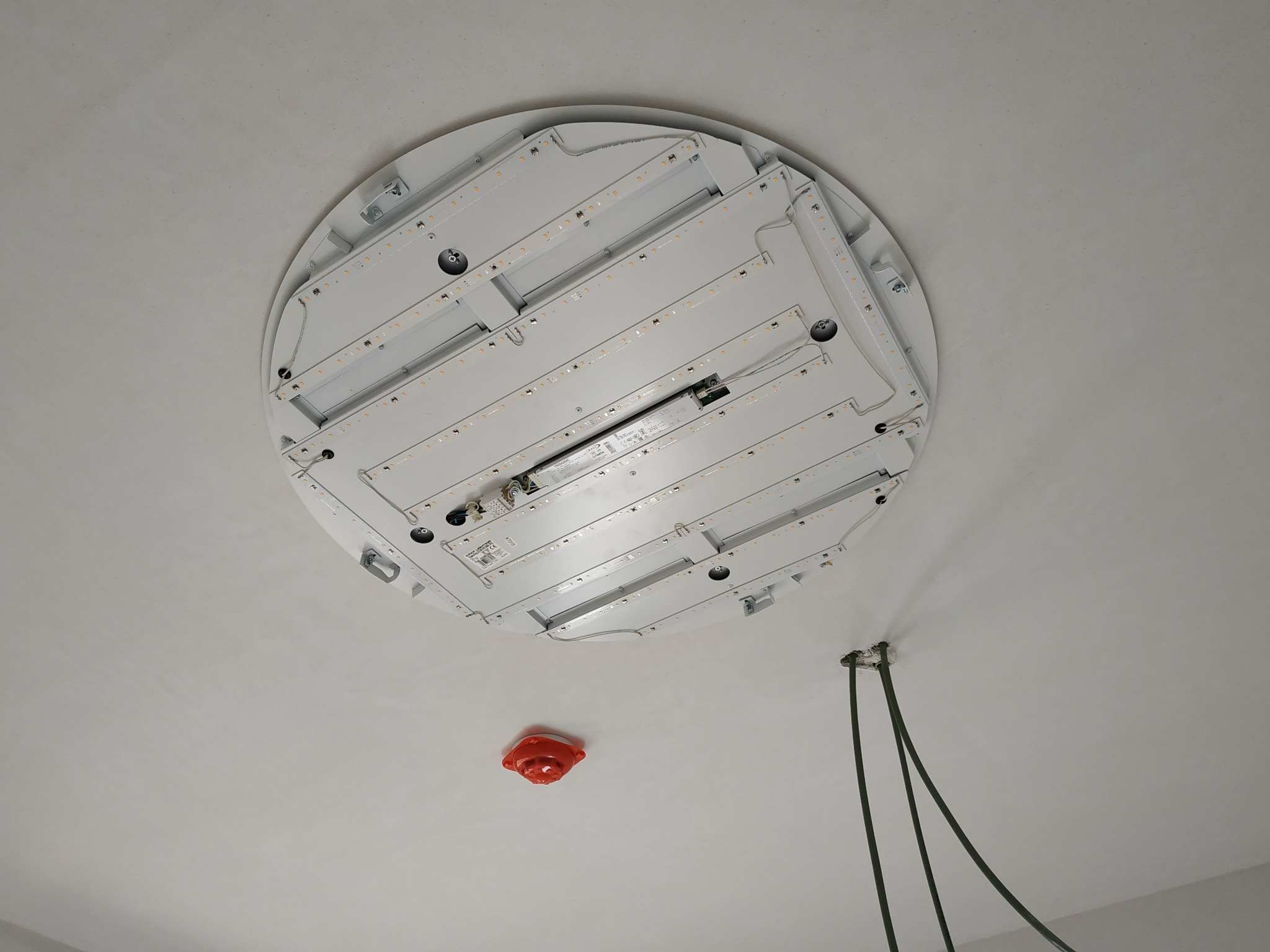 Widok od dołu na okrągłą lampę LED w trakcie montażu na białym suficie, widoczne przewody elektryczne i czujnik dymu.