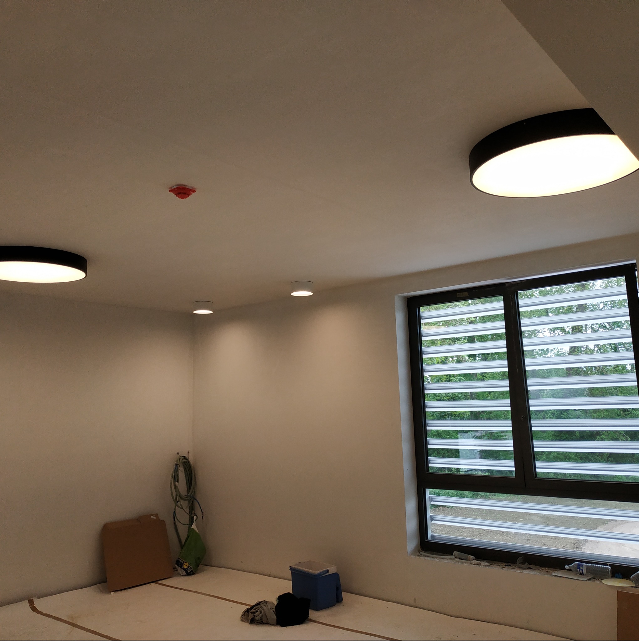 Wnętrze pokoju z trzema zamontowanymi okrągłymi lampami sufitowymi LED, czujnikiem dymu, oknem z żaluzjami i kablami elektrycznymi zwisającymi ze ściany, sugerujące prace instalacyjne w toku.