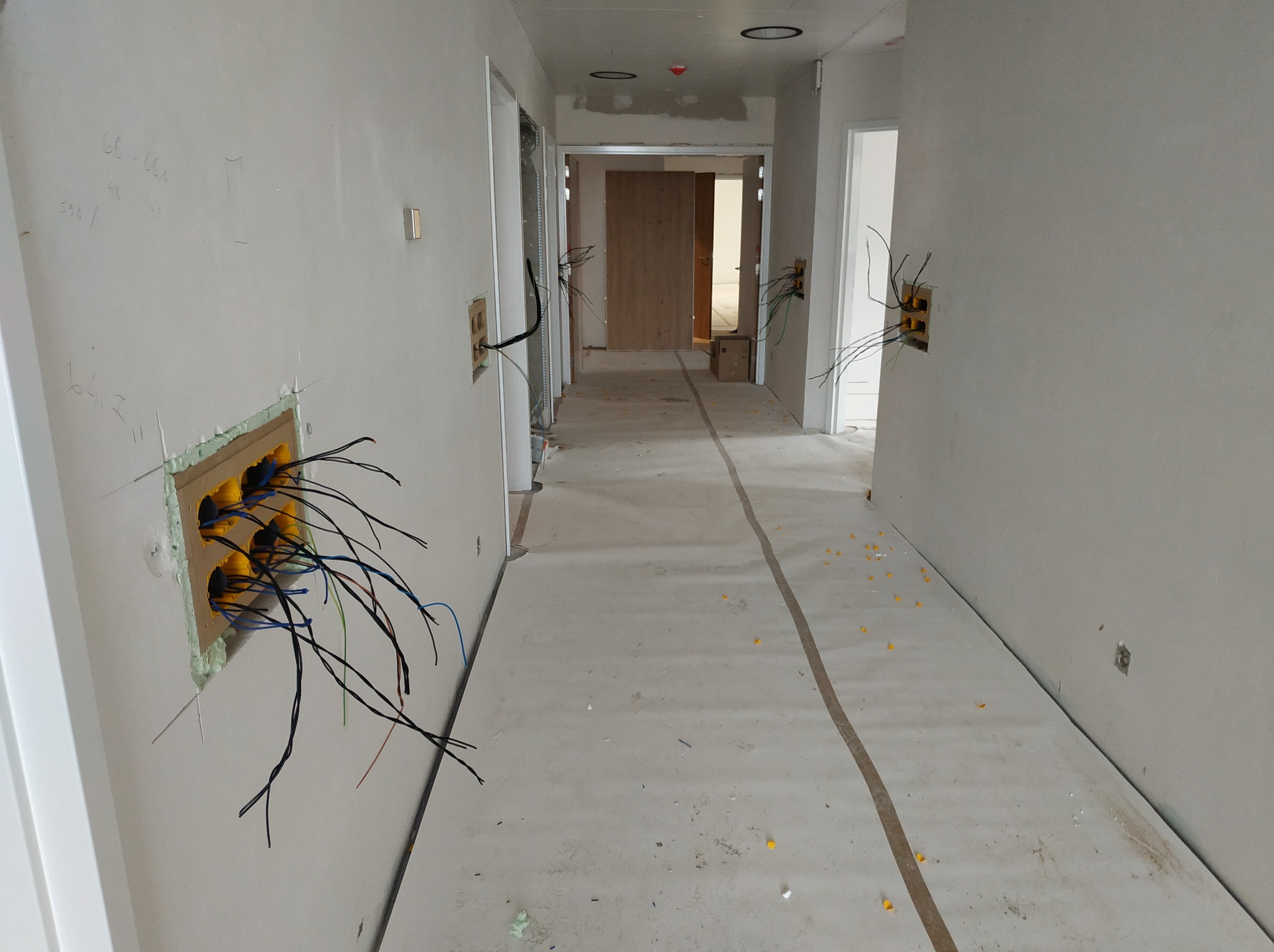 Korytarz w trakcie prac wykończeniowych, widoczne wystające przewody elektryczne z żółtymi puszkami instalacyjnymi w ścianach, zabezpieczona podłoga, surowy stan ścian.