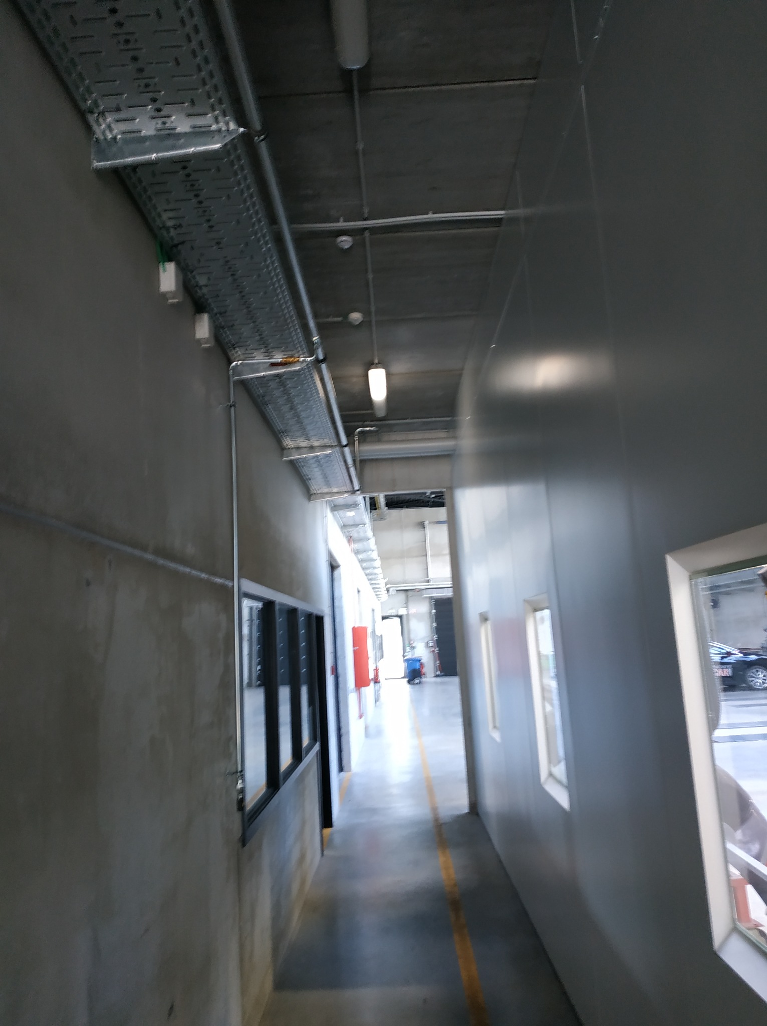 Szara, przemysłowa przestrzeń z widoczną korytką kablową pod sufitem, rurami instalacyjnymi na ścianie i oknami wychodzącymi na zewnątrz.