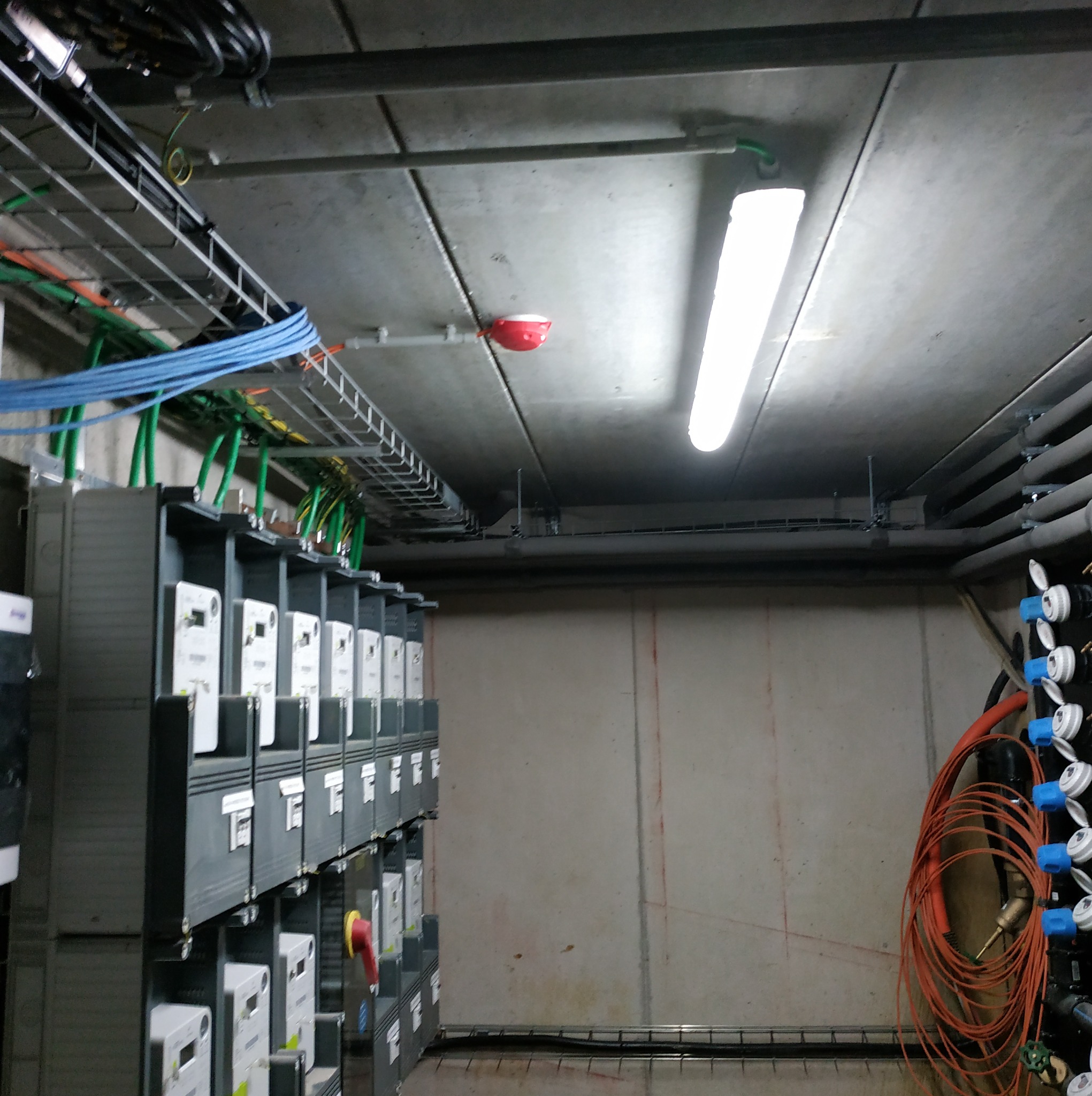 Rząd szarych szafek licznikowych z podłączonymi kablami w kolorach zielonym i niebieskim, widoczne koryta kablowe pod sufitem i oświetlenie jarzeniowe, na ścianie rury z zaworami.