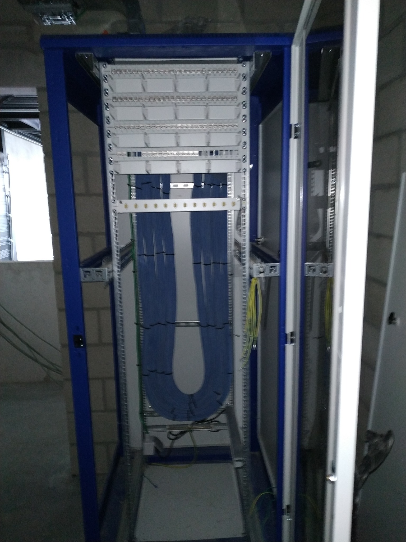 Niebieskie i zielone przewody uporządkowane w szafie rack w surowym pomieszczeniu z widoczną instalacją pod sufitem.