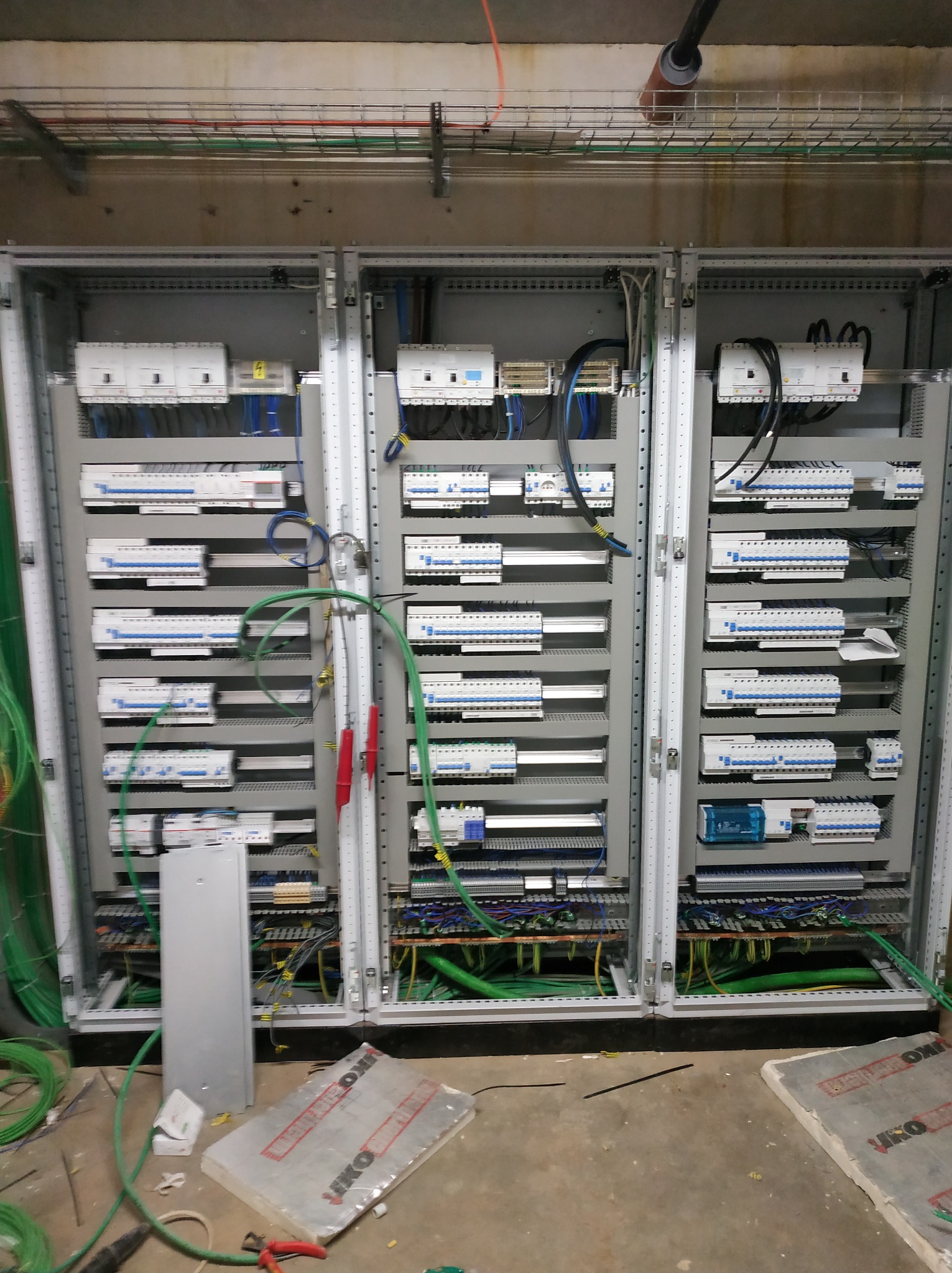 Rozdzielnia elektryczna składająca się z trzech szaf, widoczne liczne zabezpieczenia, przewody i szyny, w trakcie montażu, z elementami na podłodze.