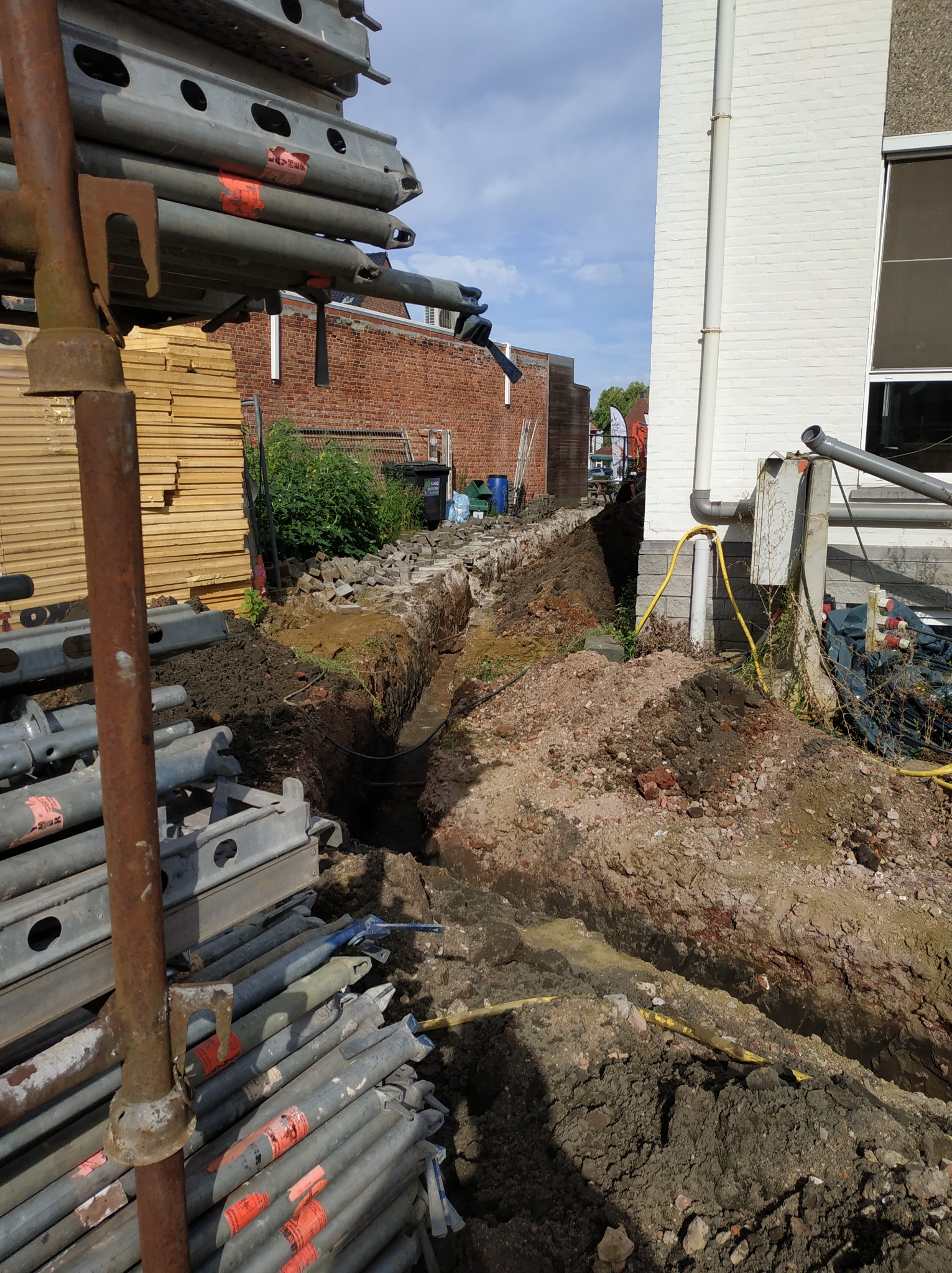 Wykop pod fundamenty budynku z widocznymi rurami instalacji elektrycznej, z hałdami ziemi i elementami rusztowania w kadrze.
