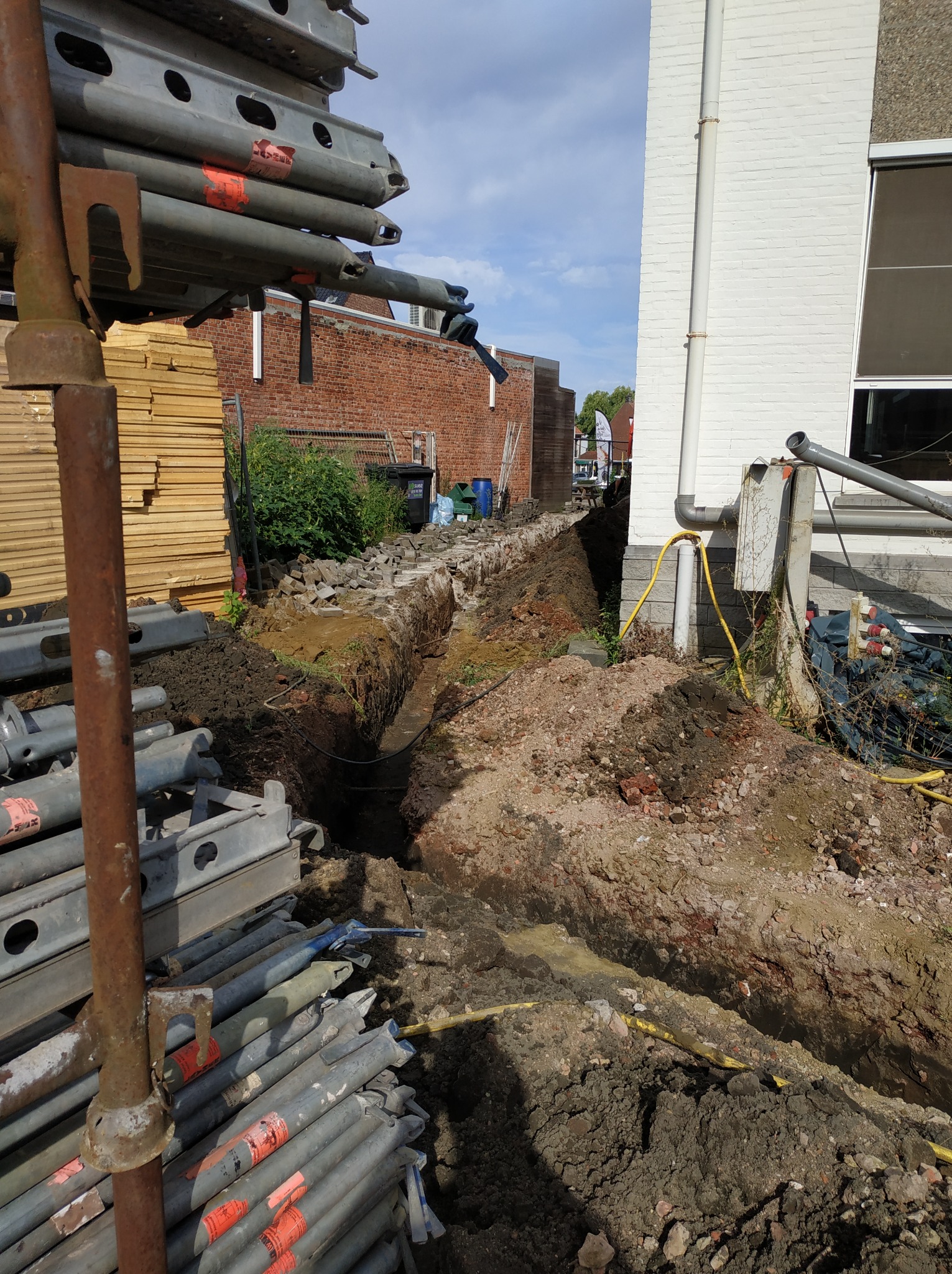 Wykop w ziemi wzdłuż ściany budynku z widocznymi rurami i przewodami, elementy rusztowania w pierwszym planie.
