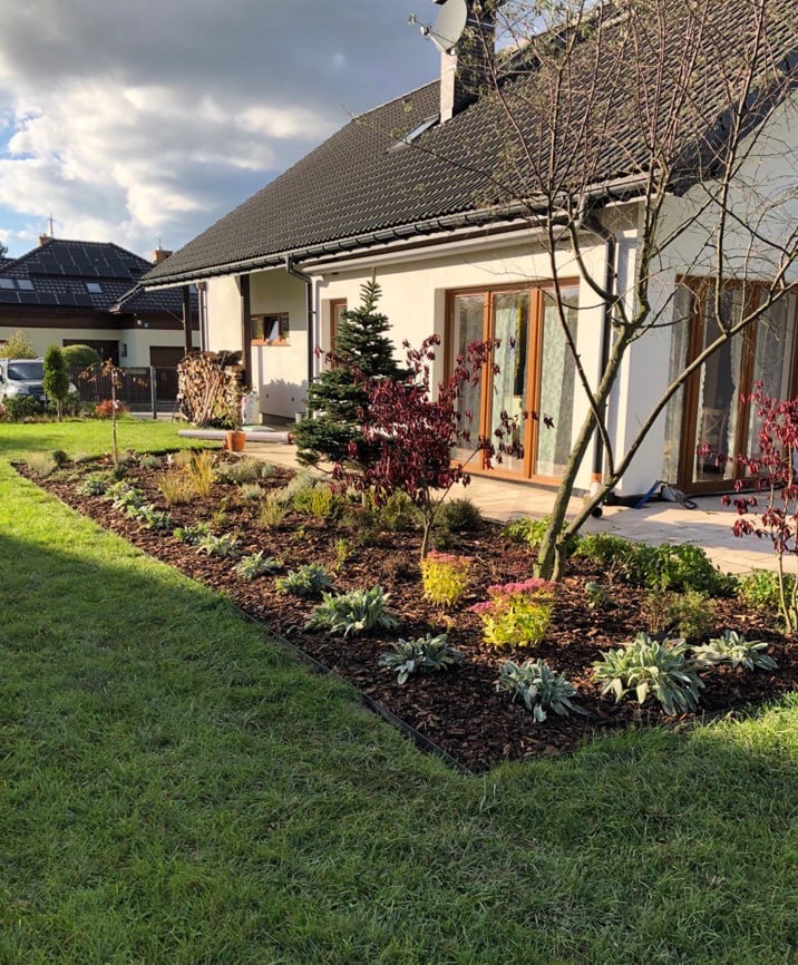 Nowoczesny ogród przydomowy z rabatą wysypaną korą, ozdobnymi krzewami i trawami, widok na dom z dużymi oknami i zadbanym trawnikiem.