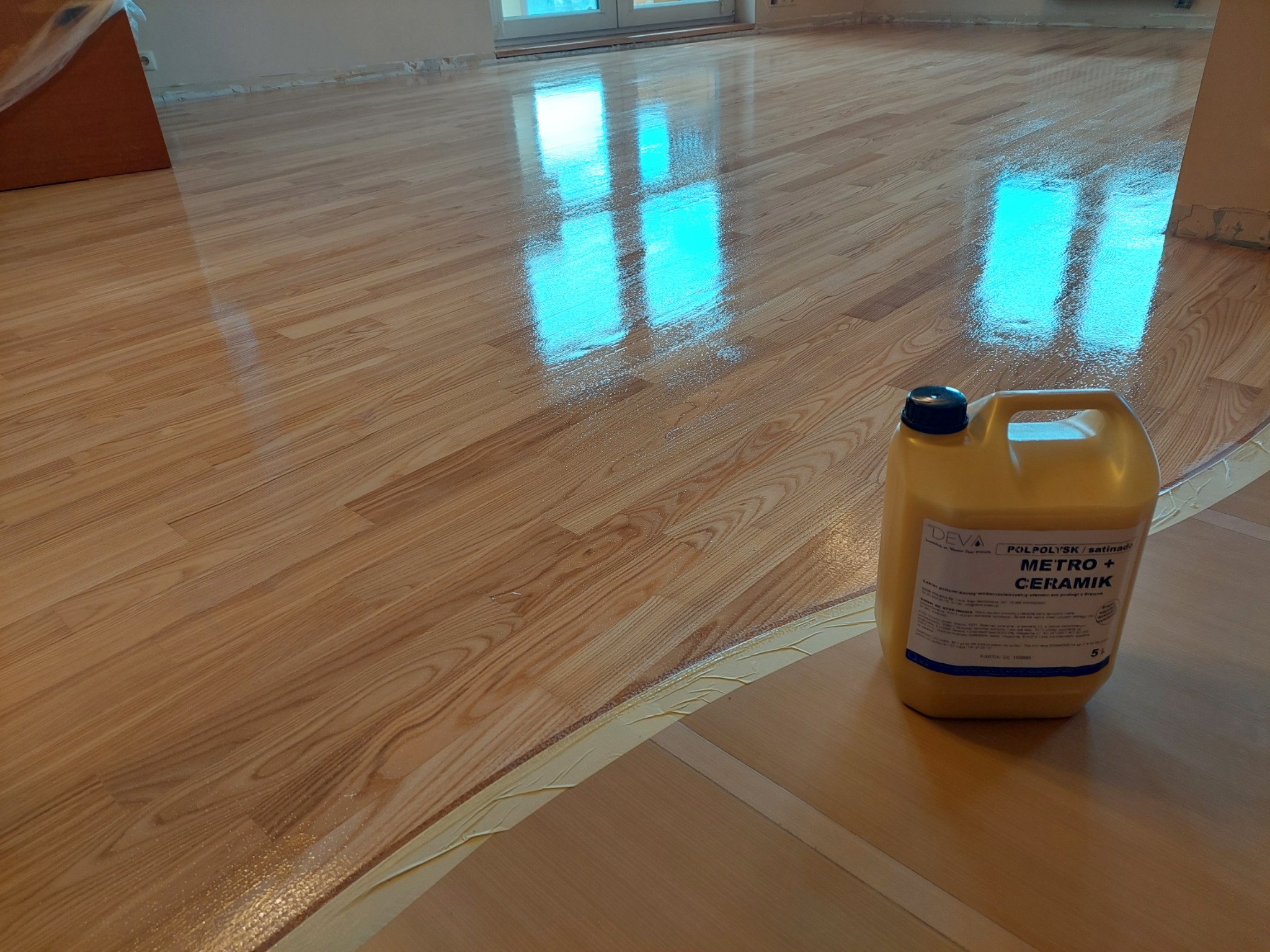 Świeżo polakierowany parkiet z widocznym odbiciem okna, na podłodze stoi kanister z lakierem POLPOLYSK Metro + CERAMIK