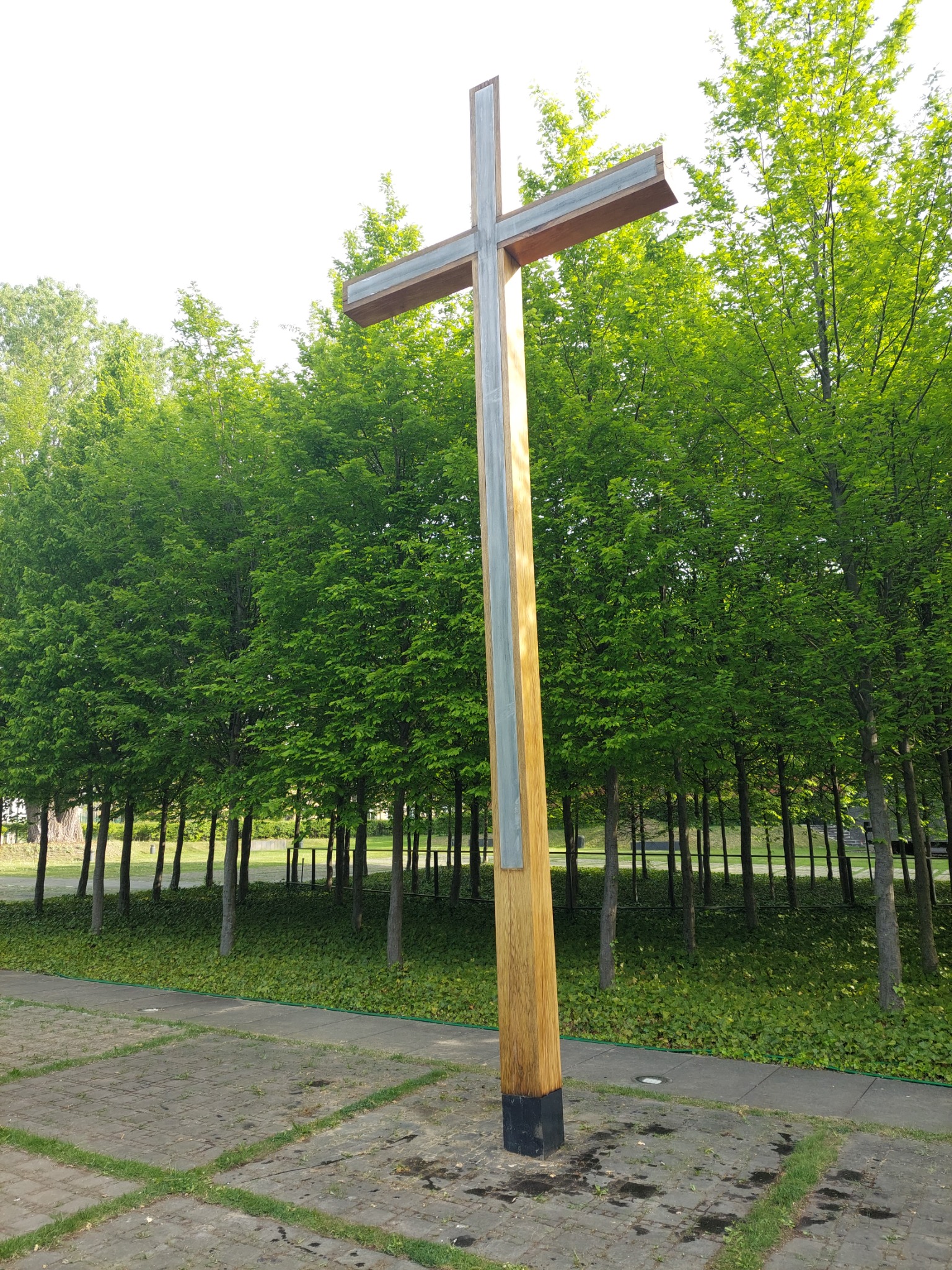 Wysoki, drewniano-metalowy krzyż na tle zielonego drzewostanu i brukowanej nawierzchni.