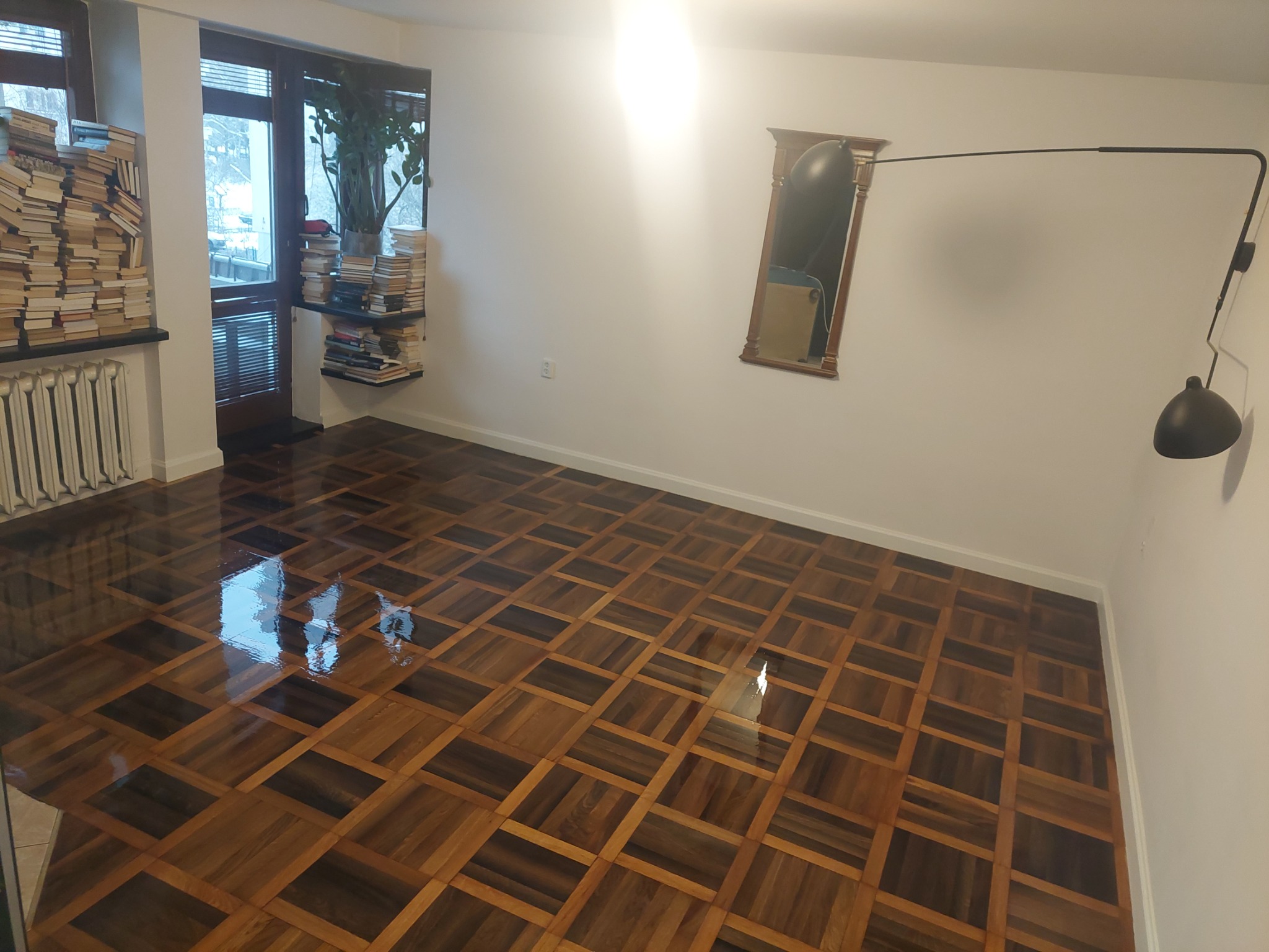 Błyszcząca, odnowiona podłoga z parkietu ułożonego w kwadraty, odbijająca światło okna w przestronnym salonie z minimalistycznym wystrojem, gdzie ściany są białe, a przy oknie stoi regał z książkami.