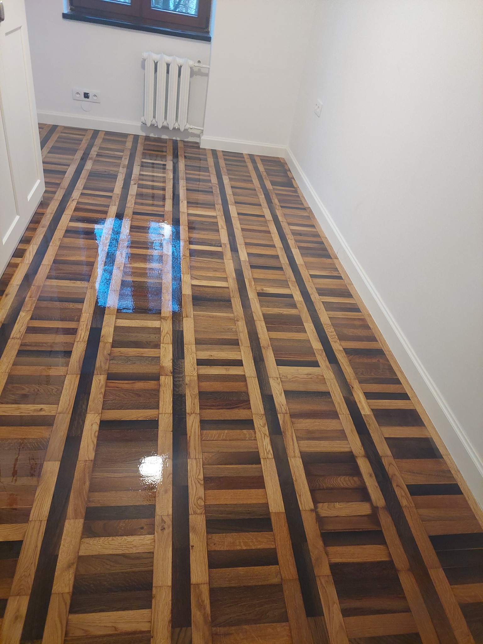 Wypolerowana, lakierowana podłoga parkietowa z jasnymi i ciemnymi elementami drewna ułożonymi w pasy, odbijająca światło z okna.