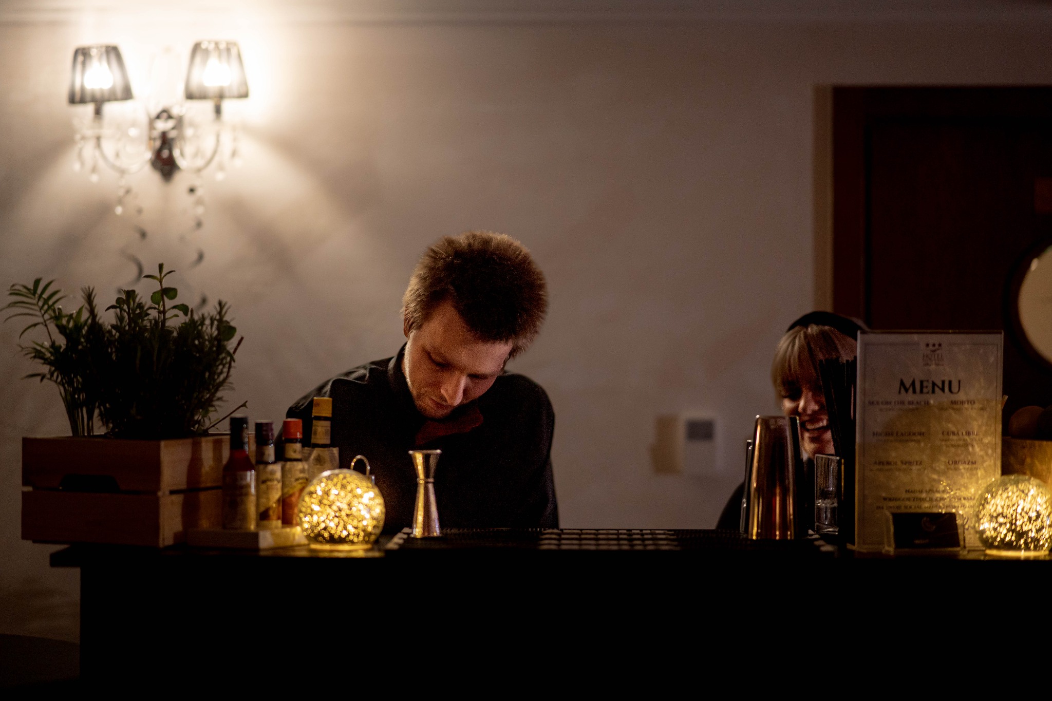 Barman przygotowuje drinki za barem, oświetlonym dekoracyjnymi kulami, w tle widoczna karta menu i uśmiechnięta kobieta.