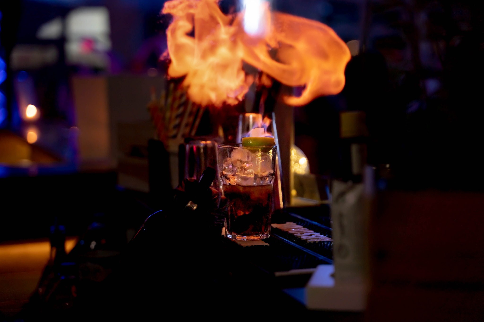 Płonący koktajl z lodem i limonką w szklance na barze, w tle butelki i inne akcesoria barmańskie.