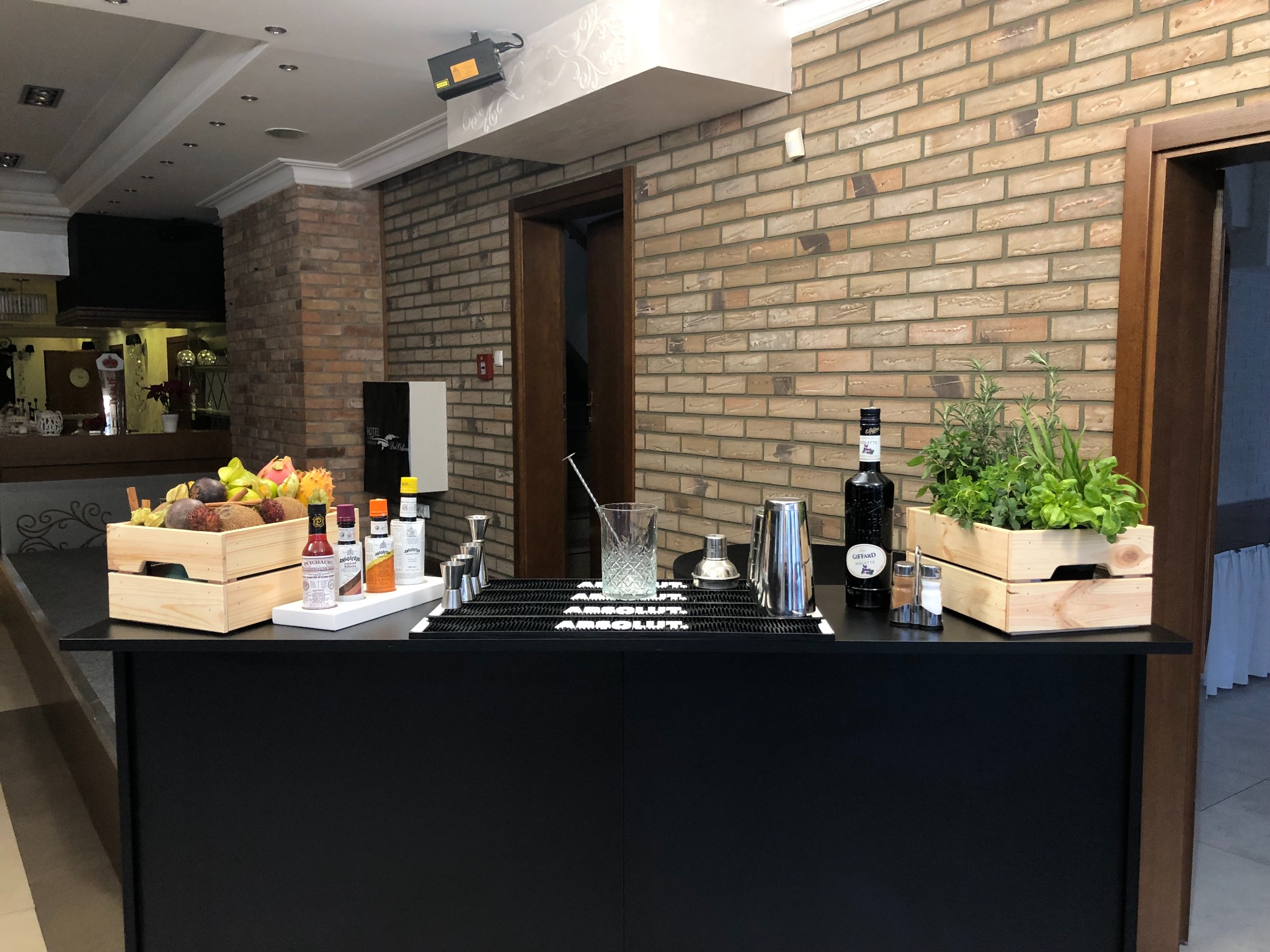 Mobilny bar z czarnym blatem, wyposażony w skrzynki z egzotycznymi owocami i ziołami, butelki z syropami, shaker, miarki i szkło do drinków, na tle ceglanej ściany.