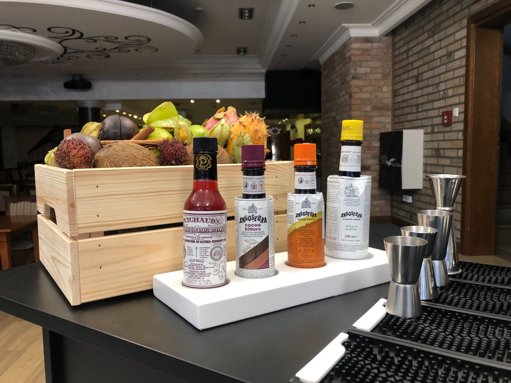 Przygotowane do drinków: skrzynka z egzotycznymi owocami i butelki z bitterami Angostura, obok miarki barmańskie, w tle ceglana ściana.