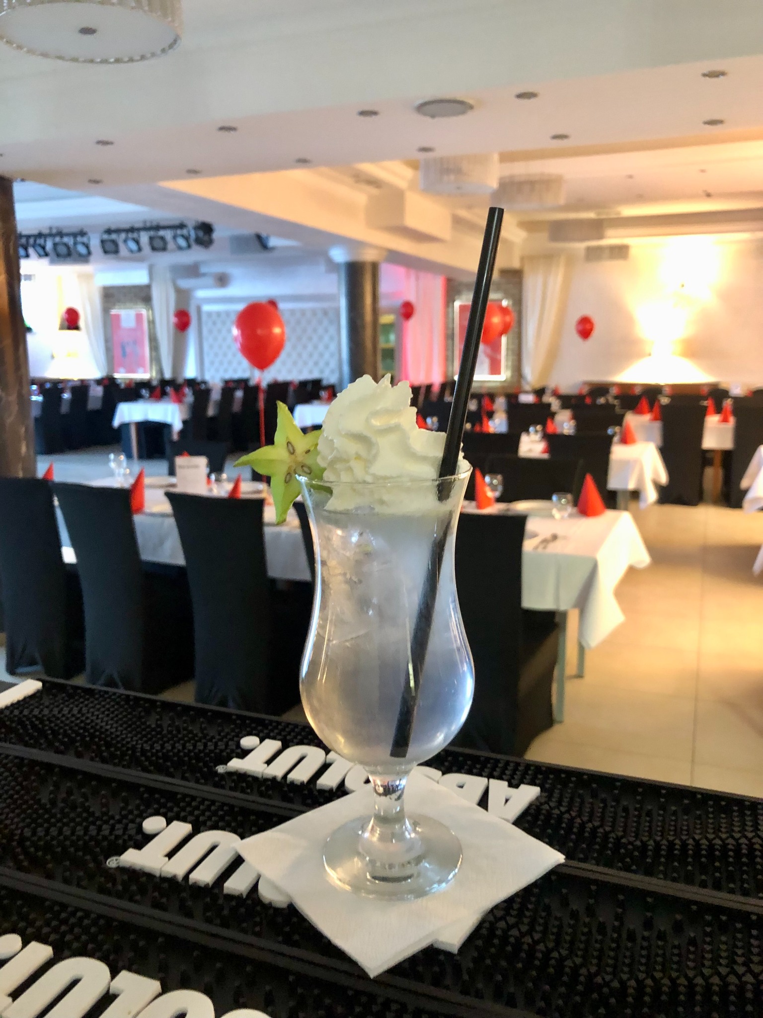 Kieliszek z białym drinkiem, bitą śmietaną i plasterkiem karamboli na barze; w tle sala bankietowa z dekoracjami i nakrytymi stołami.