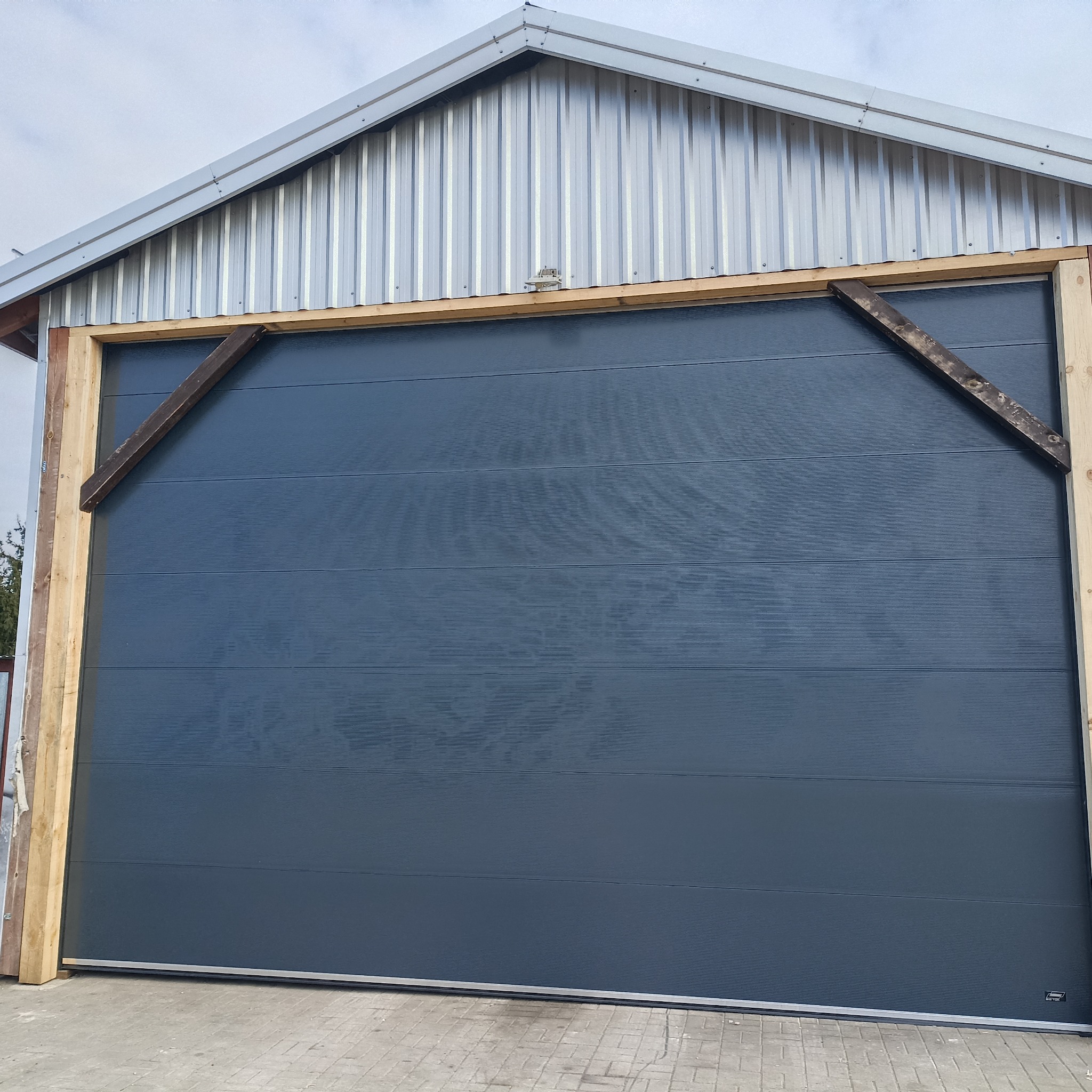 Szara segmentowa brama garażowa z drewnianą ramą montażową i metalowym dachem budynku w tle.