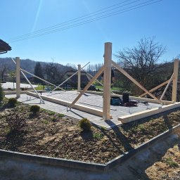 Altany drewniane Matbud - Wznoszenie drewnianej konstrukcji tarasu na kostce brukowej w ogrodzie, widoczne narzędzia i elementy konstrukcyjne, w tle dom i drzewa.