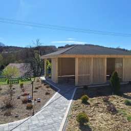 Altany drewniane Matbud - Nowoczesna altana z drewnianymi lamelami i ciemnym dachem na tle zieleni. Płytkowy chodnik prowadzi do wejścia. Widoczny fragment osoby w odblaskowej kamizelce.