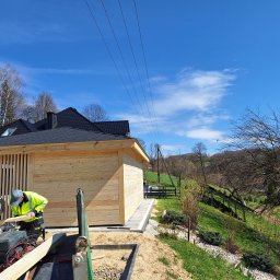 Altany drewniane Matbud - Pracownik w odblaskowej kamizelce tnie drewno piłą mechaniczną na tle drewnianej altany z czarnym dachem i błękitnego nieba. Widoczne narzędzia i otoczenie w trakcie budowy.