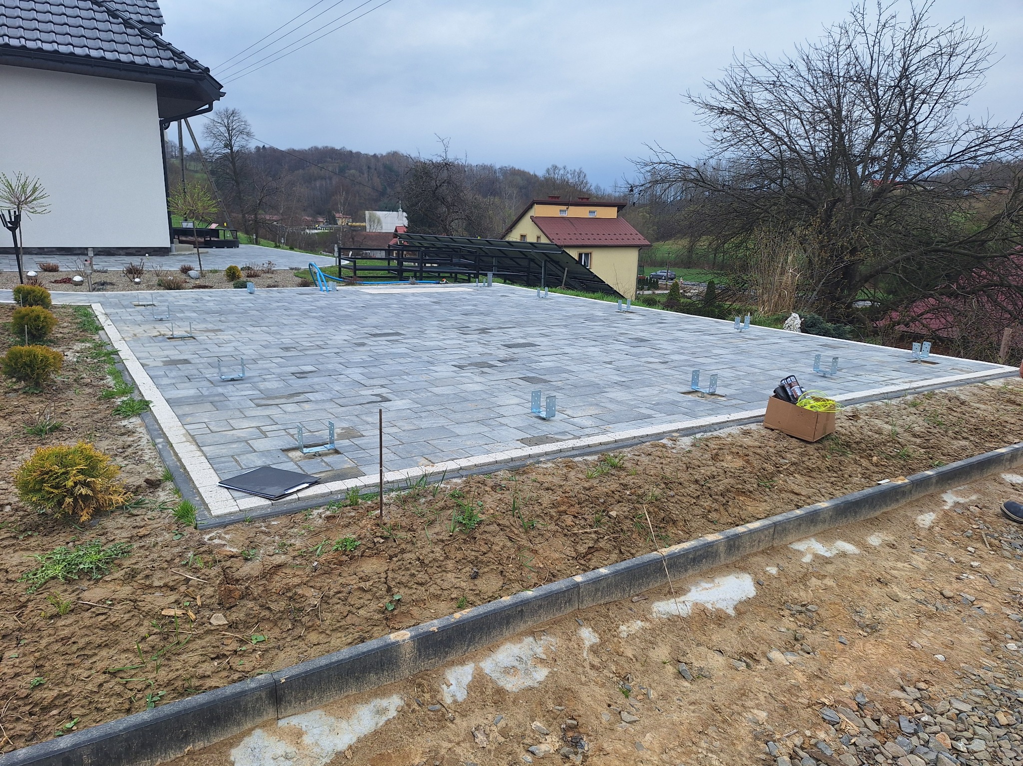 Przygotowany podkład z kostki brukowej pod budowę altany z metalowymi wspornikami, widok z góry na tle zabudowań i drzew w pochmurny dzień.