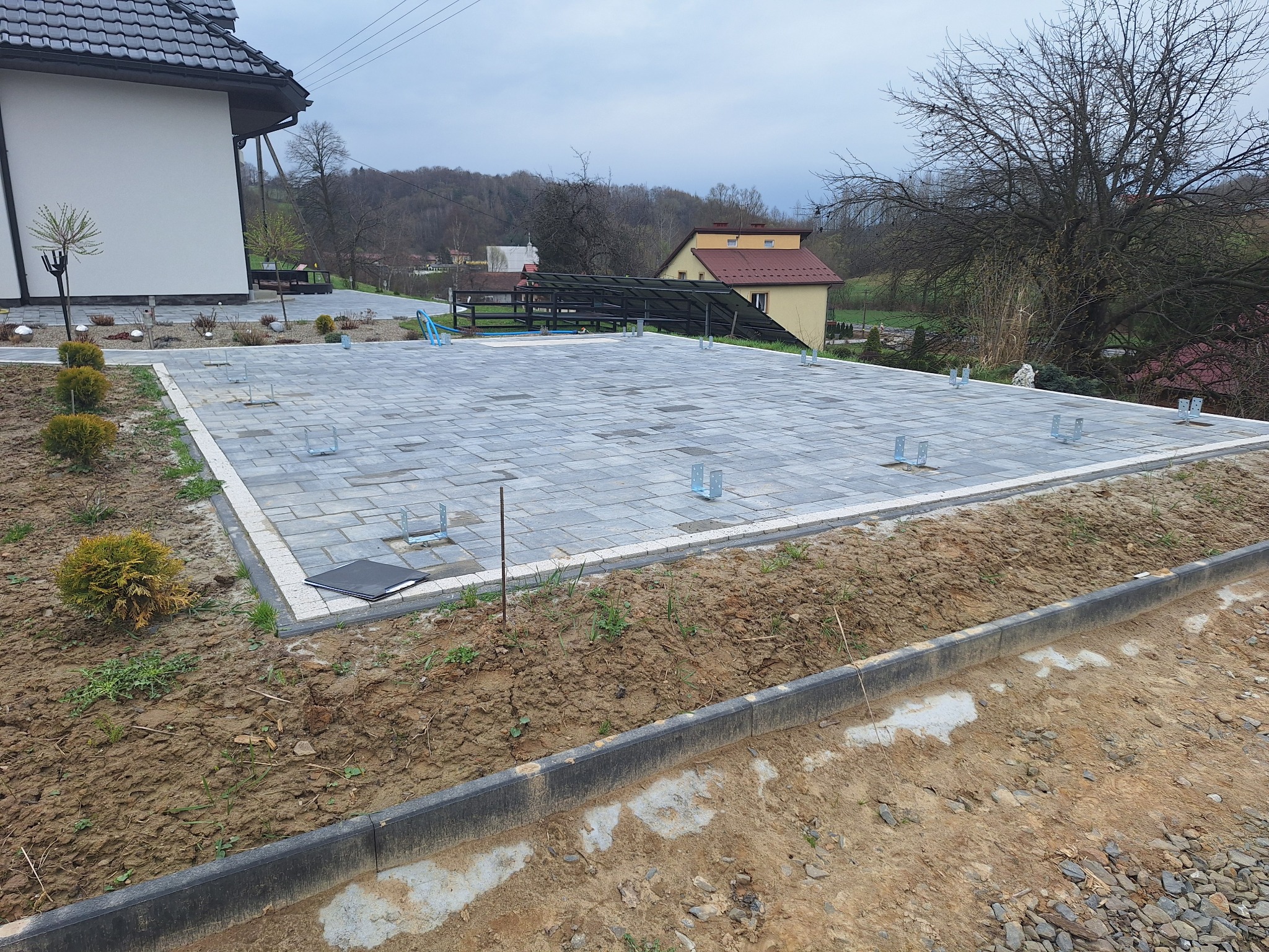 Ułożona kostka brukowa na przygotowanym podłożu z wystającymi metalowymi elementami, prawdopodobnie pod przyszłą konstrukcję tarasu. Widoczne obrzeża i przygotowane rabaty.