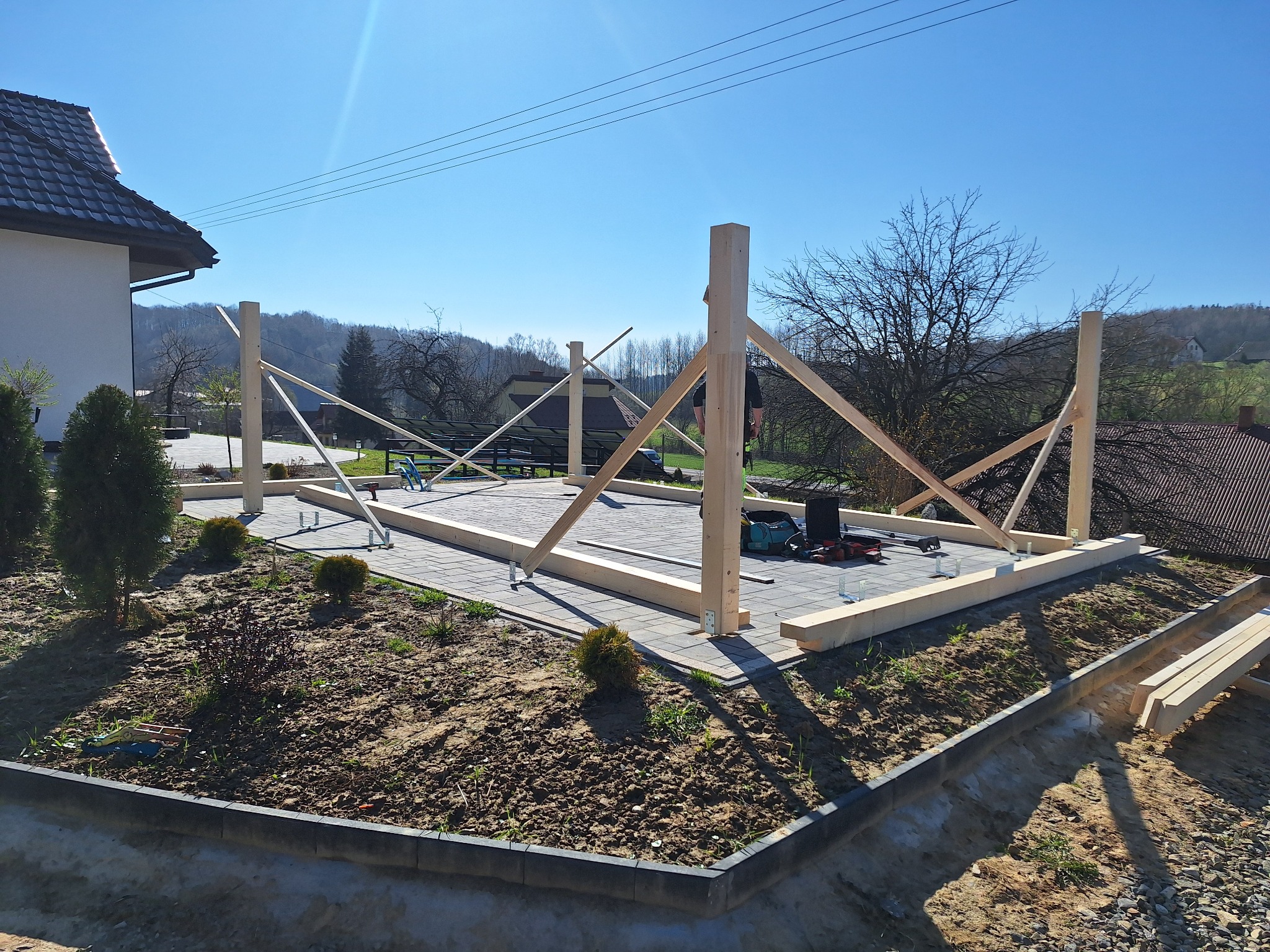 Wznoszenie drewnianej konstrukcji tarasu na kostce brukowej w ogrodzie, widoczne narzędzia i elementy konstrukcyjne, w tle dom i drzewa.