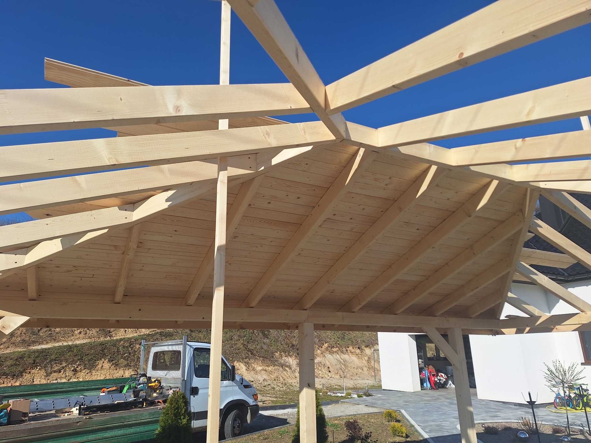 Drewniana konstrukcja zadaszenia w trakcie budowy, widok od spodu na deski dachu i jasne niebo. Na dole narzędzia i samochód dostawczy.