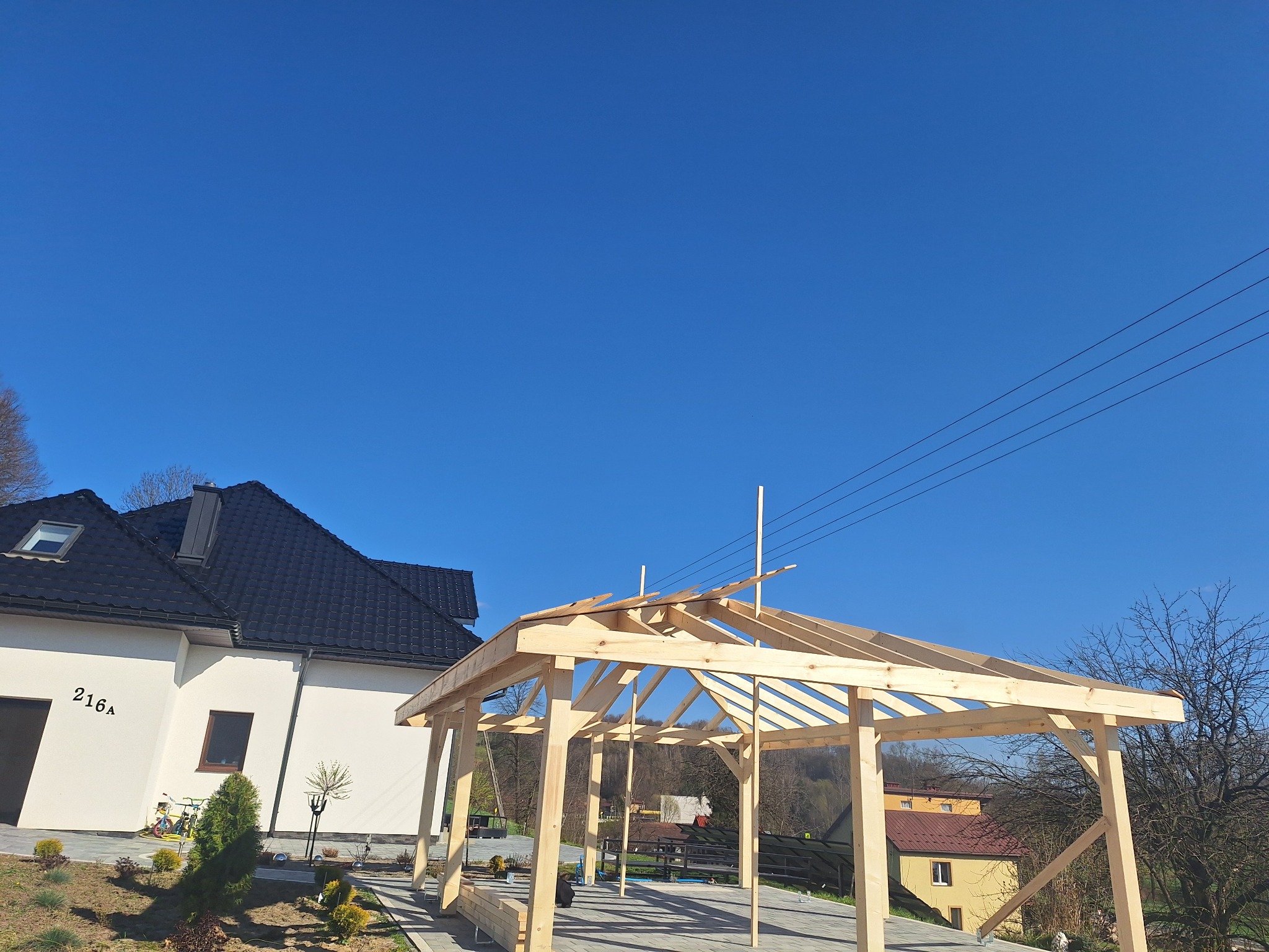 Wznoszona drewniana konstrukcja altany na tle błękitnego nieba i domu z ciemnym dachem. Widoczne elementy szkieletu i podłoże z kostki brukowej. Jasne drewno konstrukcji.