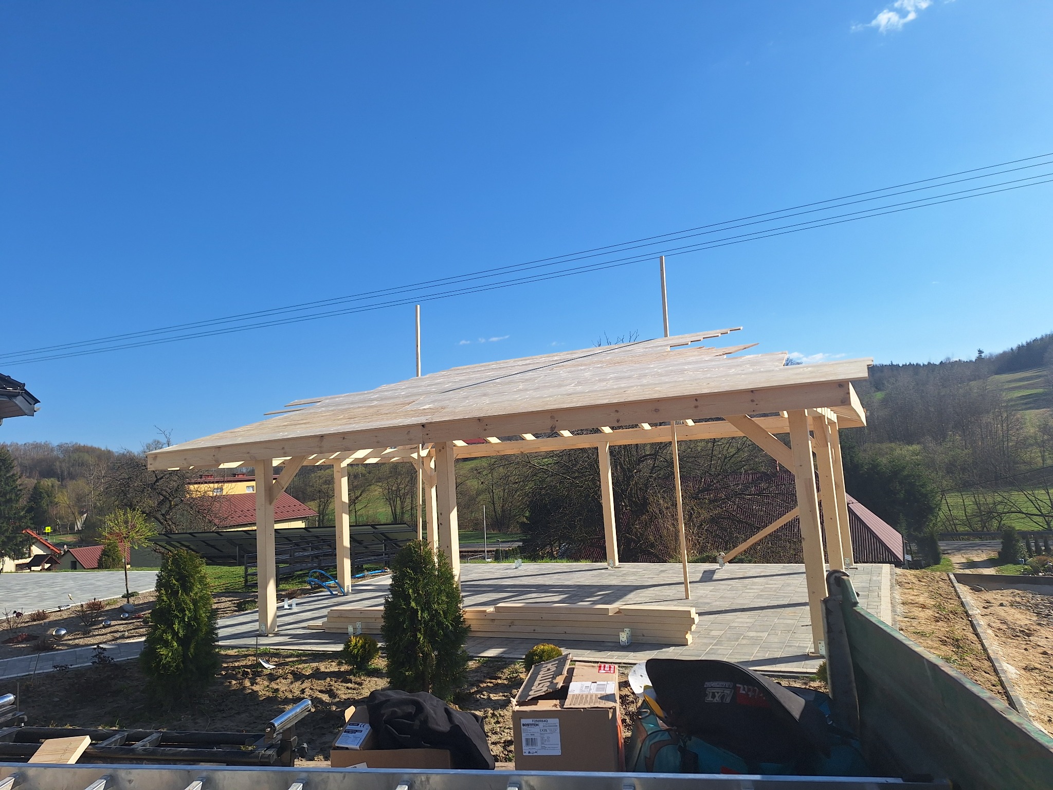 Wznoszenie drewnianej pergoli na posesji. Widoczna konstrukcja z jasnego drewna, częściowo zadaszona, z elementami montażowymi. W tle domy i zieleń.